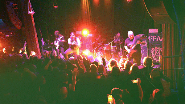 A live band performing on stage at a concert with colorful lighting, and a crowd of fans watching and taking photos.