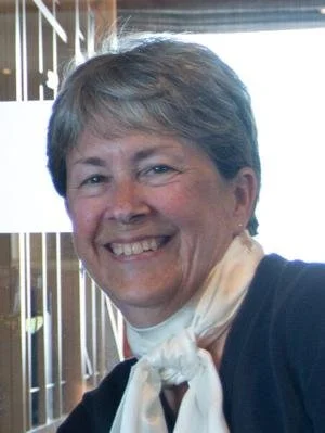 Close-up of a smiling woman with short gray hair, wearing a black top and a white scarf, sitting near a window with wooden blinds.