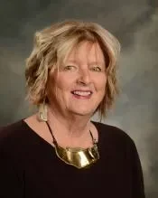 A portrait of a woman with short blond hair smiling, wearing a dark top and a large gold necklace against a gray background.