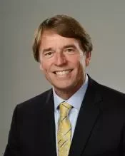 Portrait of a man with light brown hair, wearing a dark suit, white shirt, and a yellow patterned tie, smiling against a plain gray background.