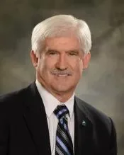 Portrait of an older man with white hair, wearing a black suit, white shirt, and striped tie, smiling at the camera.