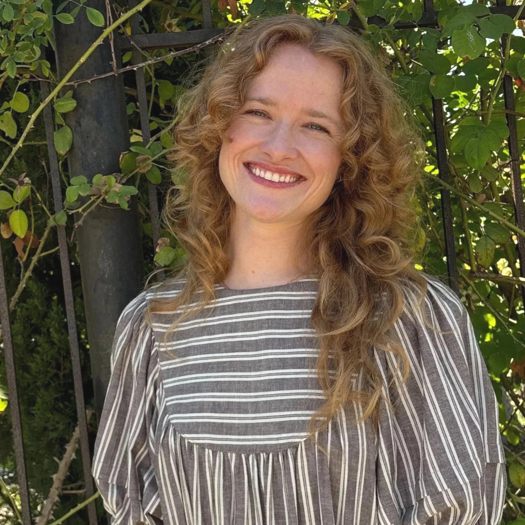 Smiling woman with long, curly red hair wearing a striped shirt, standing outdoors in front of a black metal fence and green foliage.