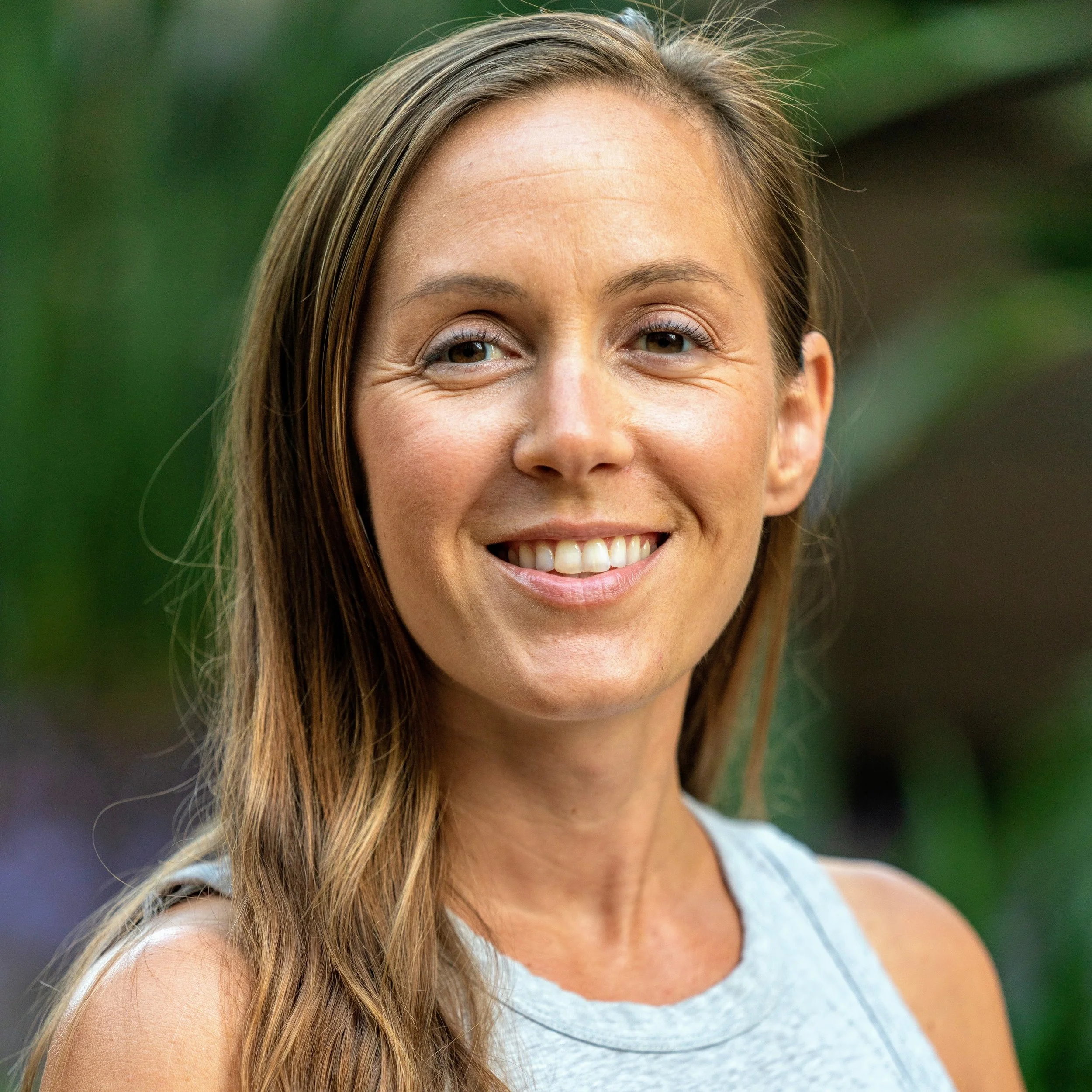 Close-up of a smiling woman with long straight brown hair, wearing a sleeveless gray top, outdoors with green foliage in the background.