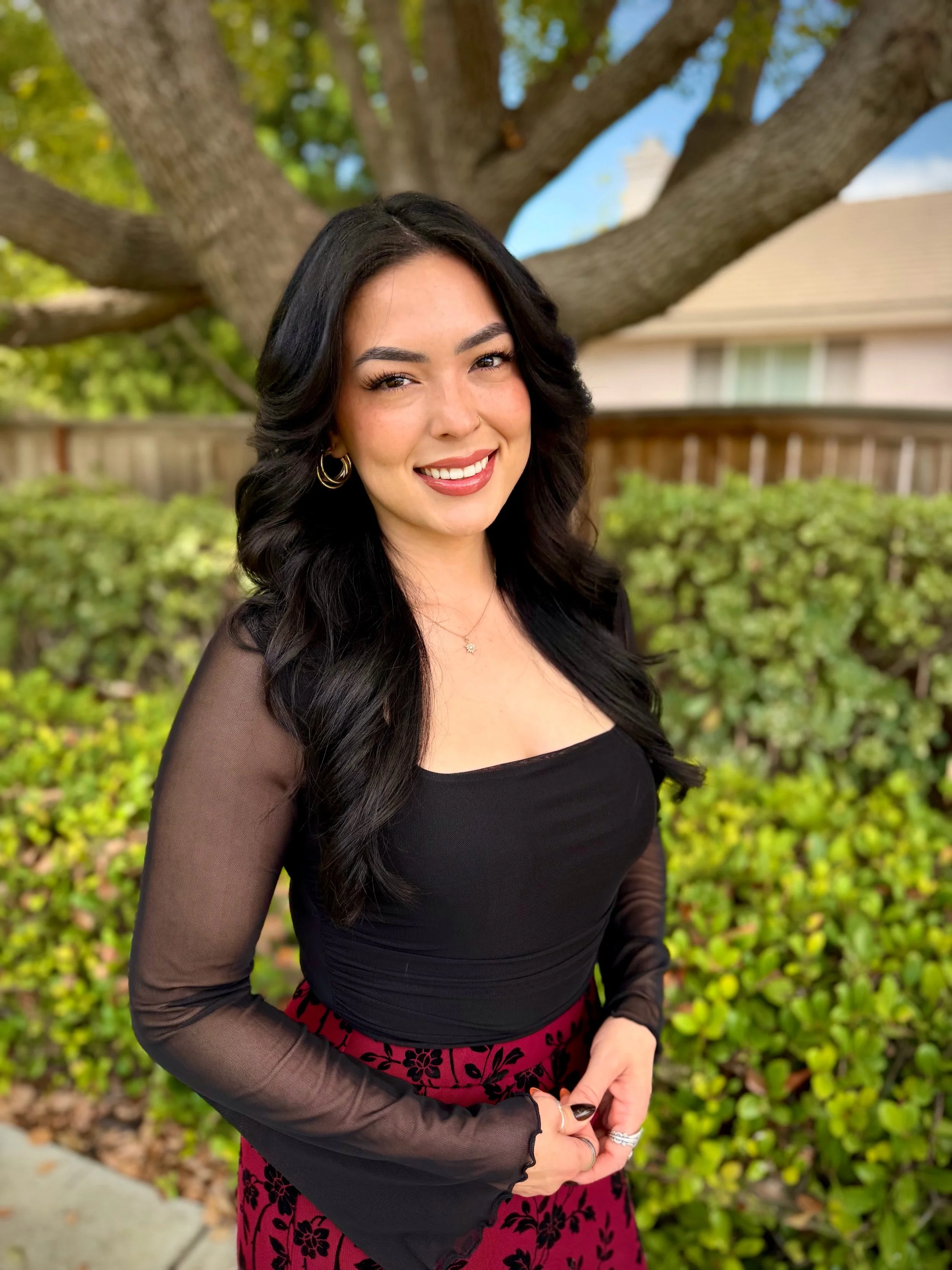 A woman with long black hair wearing a black top with sheer sleeves and a red skirt with black floral patterns, standing outdoors in front of a large tree and greenery, smiling at the camera.