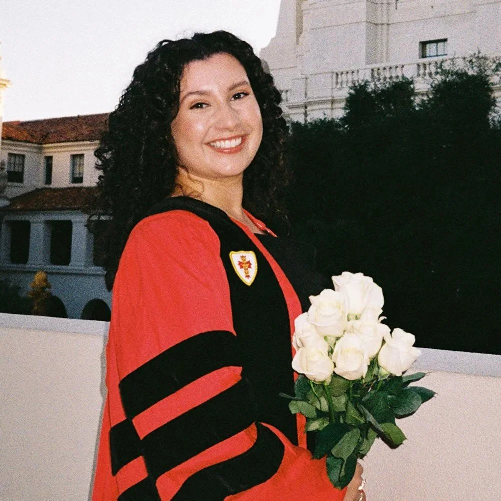 A young woman with curly dark hair smiling and holding a bouquet of white roses, wearing a black and red academic gown with a crest, standing outdoors in front of a building and trees.