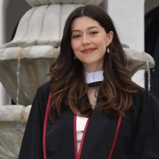 Young woman with long brown hair wearing graduation gown and cap, standing in front of a fountain.