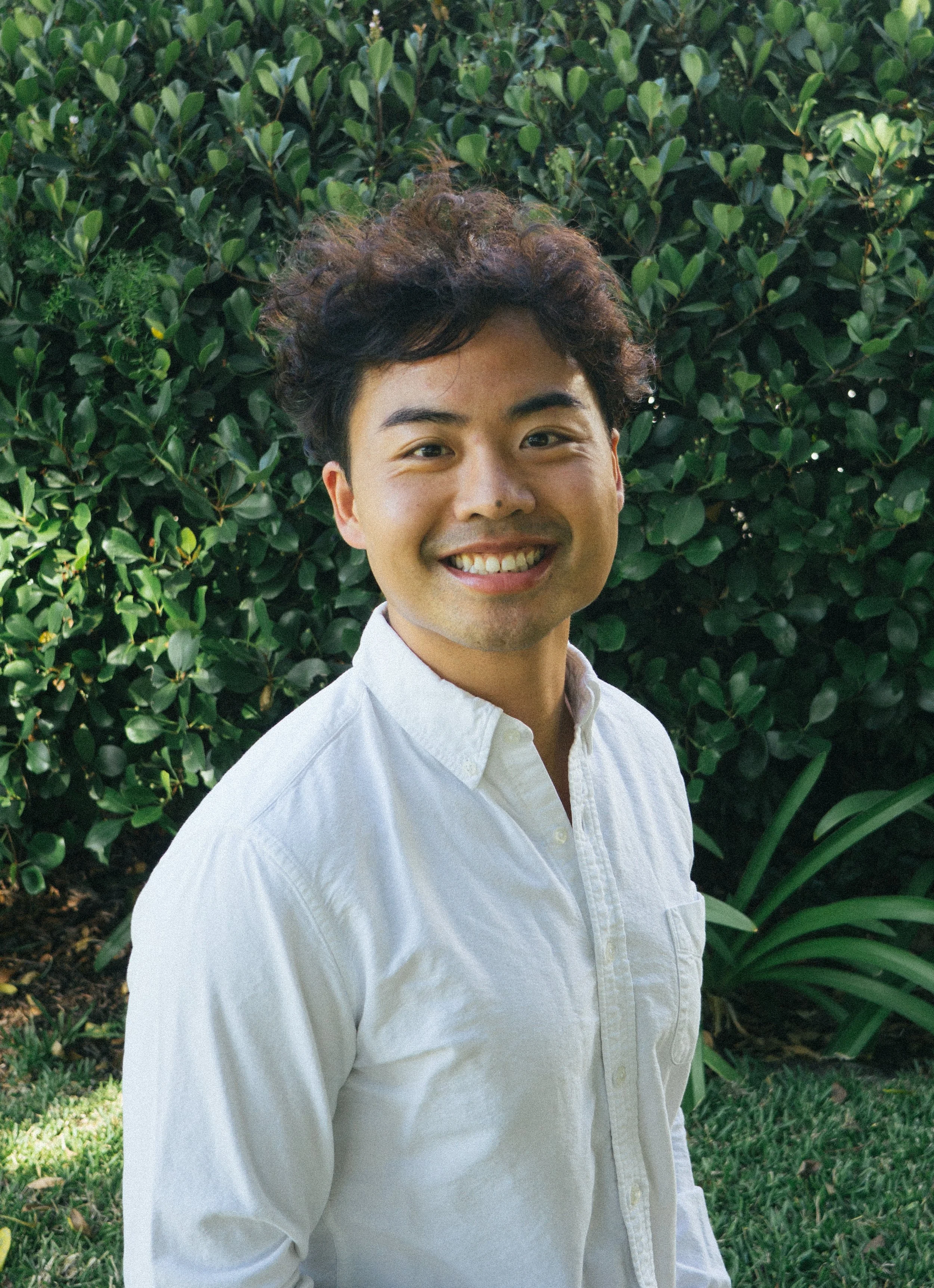 A smiling young man with curly dark hair wearing a white shirt, standing outdoors in front of green foliage.