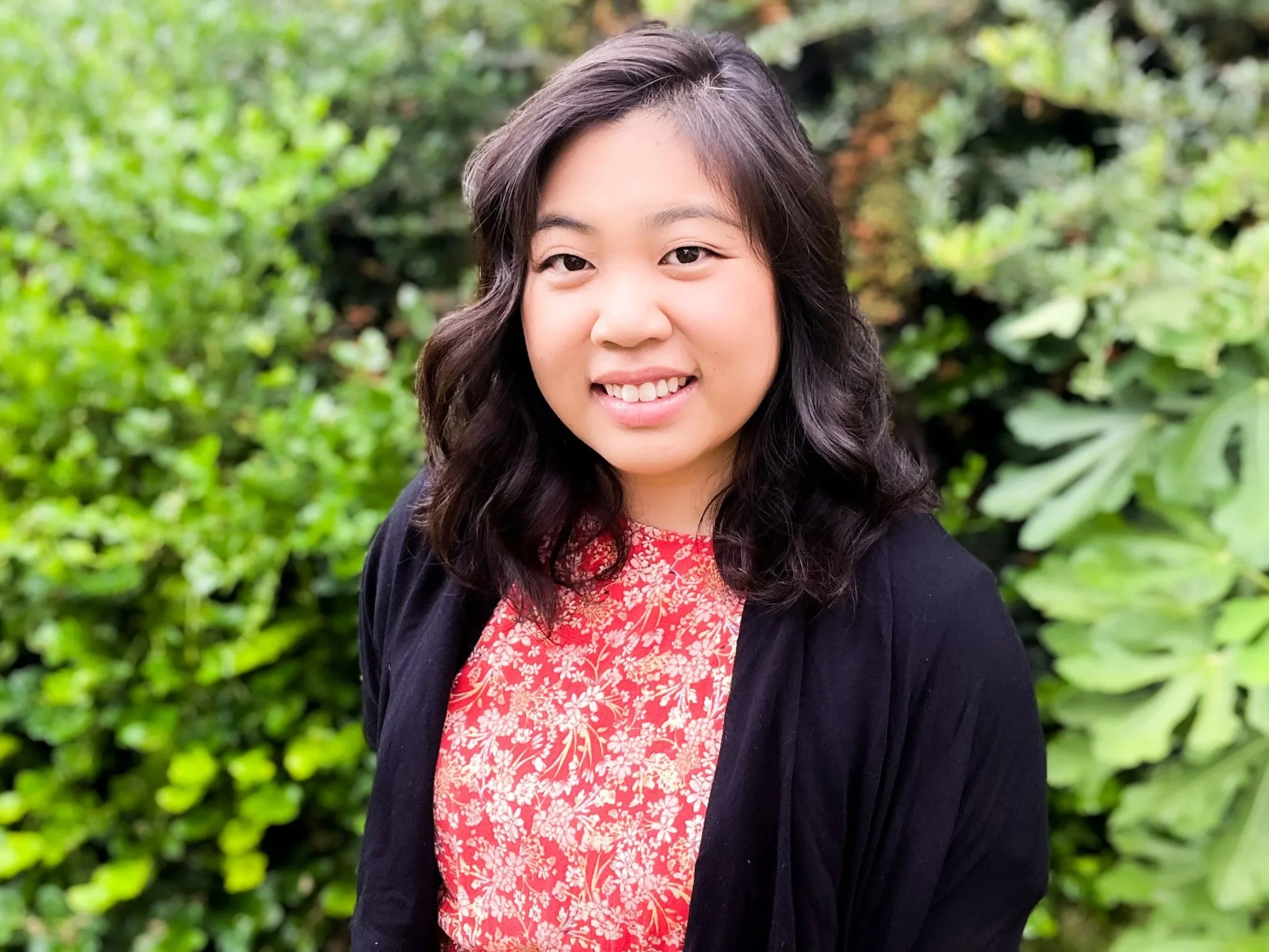 A young woman with wavy dark hair, smiling, wearing a red floral blouse and black jacket, standing outdoors in front of green foliage.