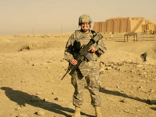 A soldier in camouflage uniform standing in a desert landscape, smiling and holding a rifle.
