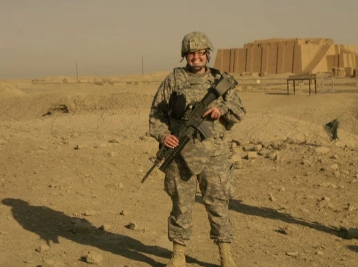 A soldier in camouflage uniform standing in a desert landscape, holding a rifle, with a large mud-brick building in the background.