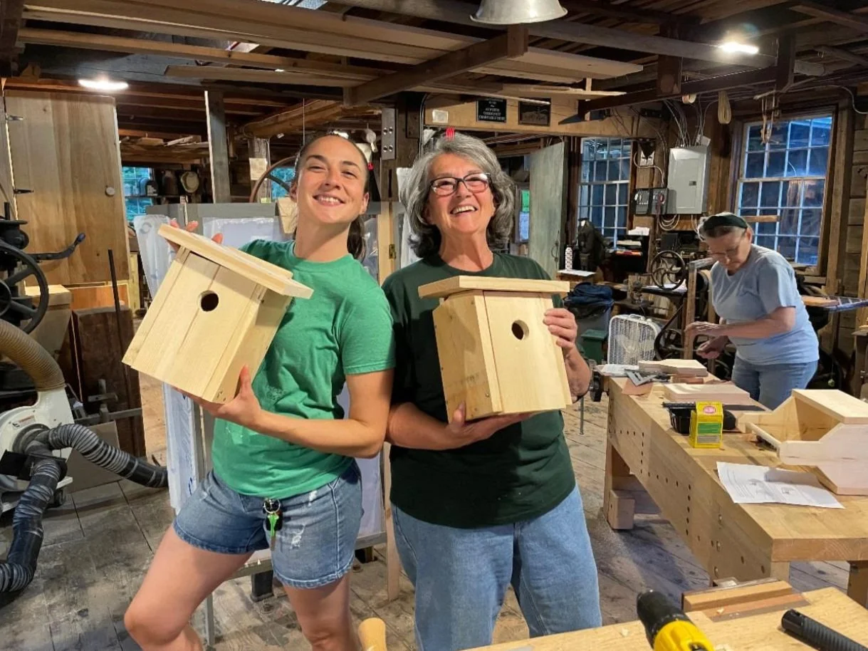 Two people holding wooden birdhouses and smiling with woodworking tools in background