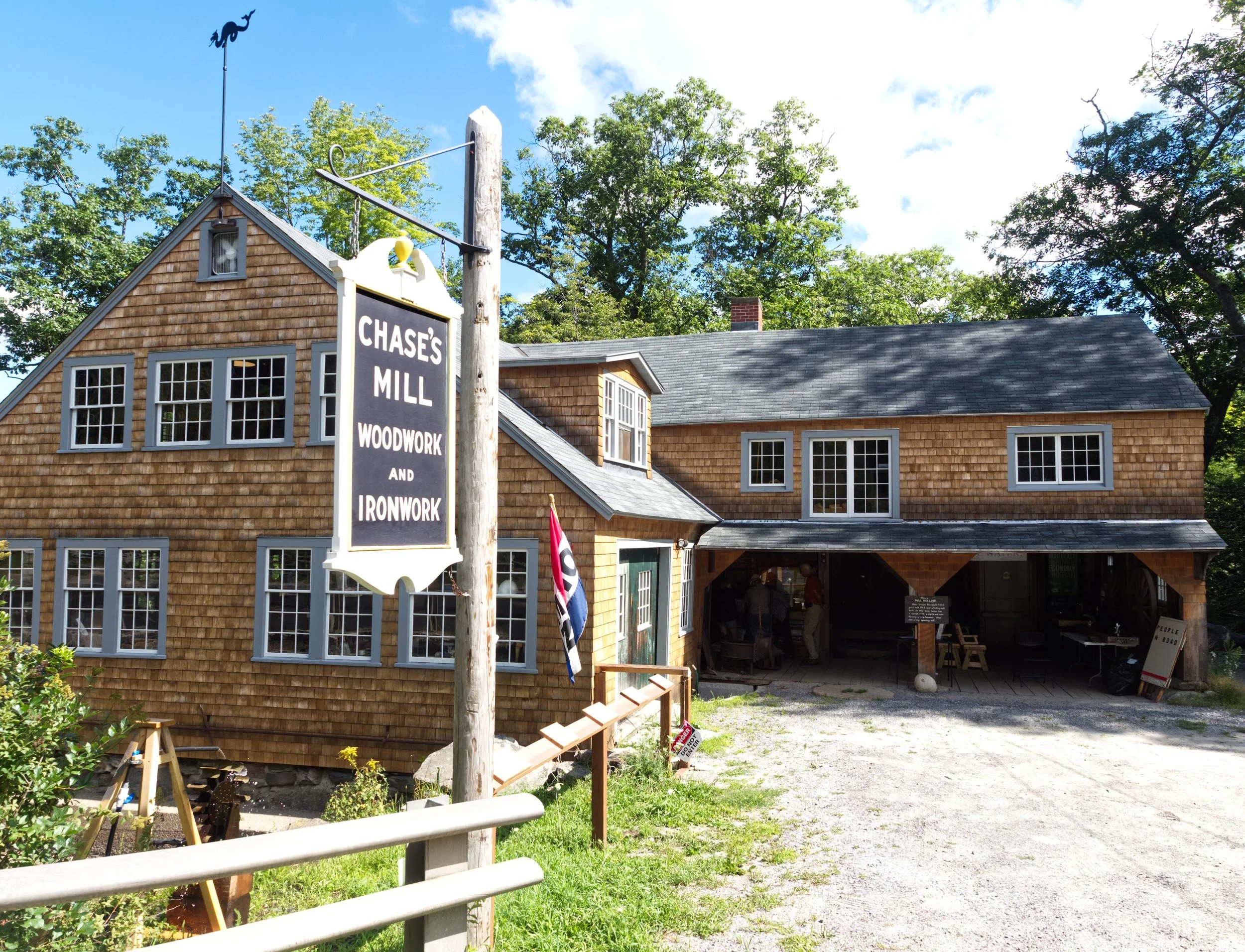 Chase's Mill building with sign in front that says "CHASE'S MILL WOODWORK AND IRONWORK." Two-story building is covered with unpainted wood shingles. Blue sky and trees in background.