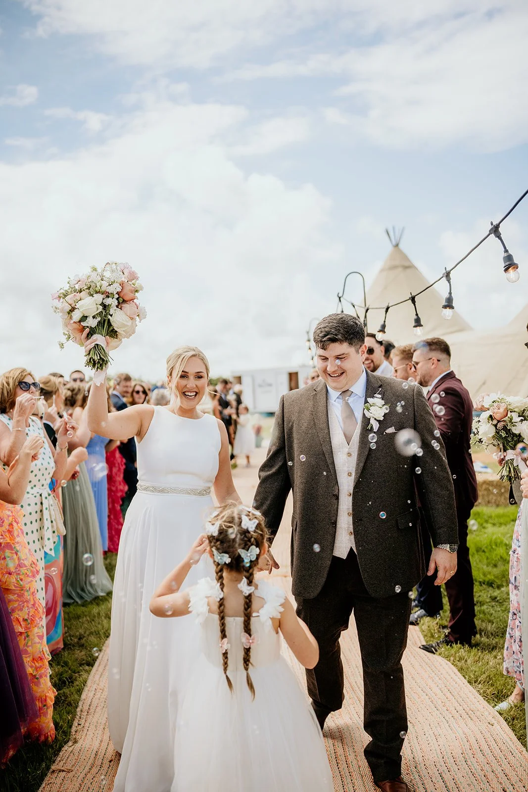 A joyful wedding celebration outdoors with a bride in a white dress and a groom in a suit, surrounded by guests. The bride is holding a bouquet and raising her hand, while the groom looks at a young flower girl with braided hair. There are tents, str