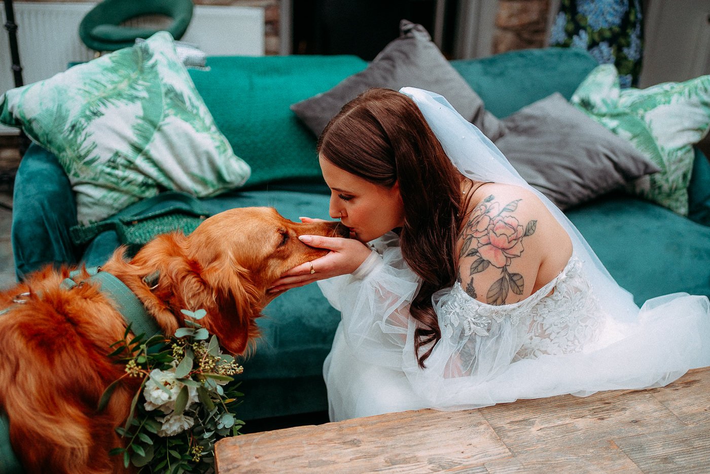 A woman in a wedding dress with a floral tattoo on her shoulder is kissing and holding her dog's face. The dog is wearing a collar and a floral wreath around its neck, sitting on a wooden table in front of a green sofa with patterned cushions.