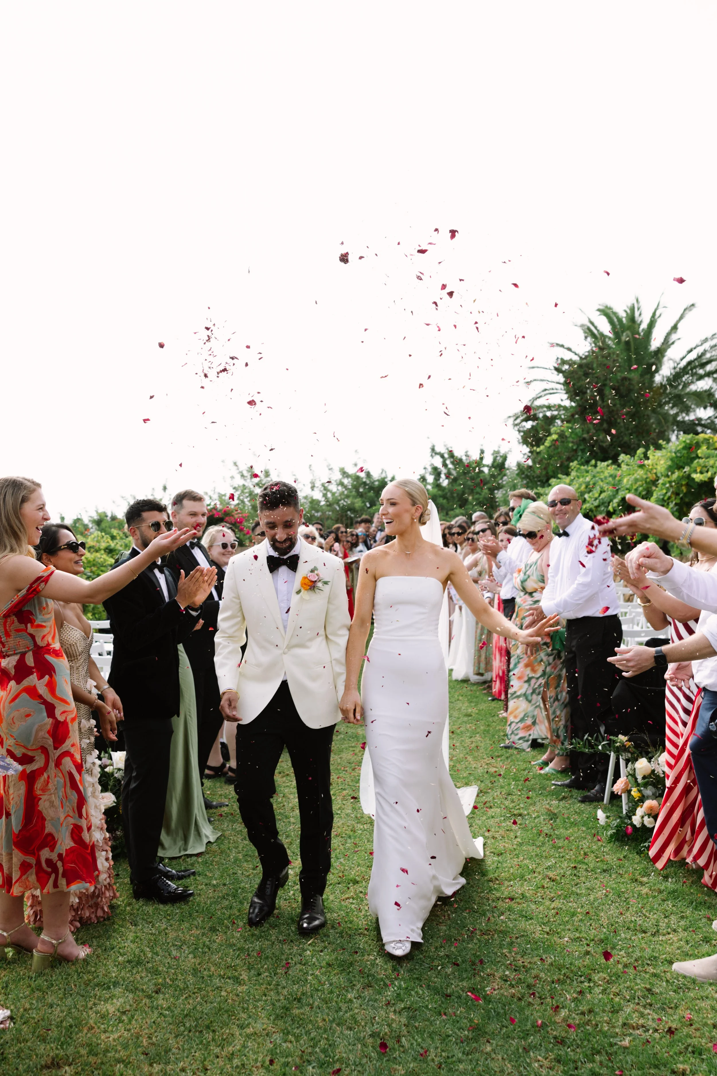 A bride and groom walking hand in hand through a celebratory crowd at their wedding, with confetti falling around them outdoors on a grassy area surrounded by trees.