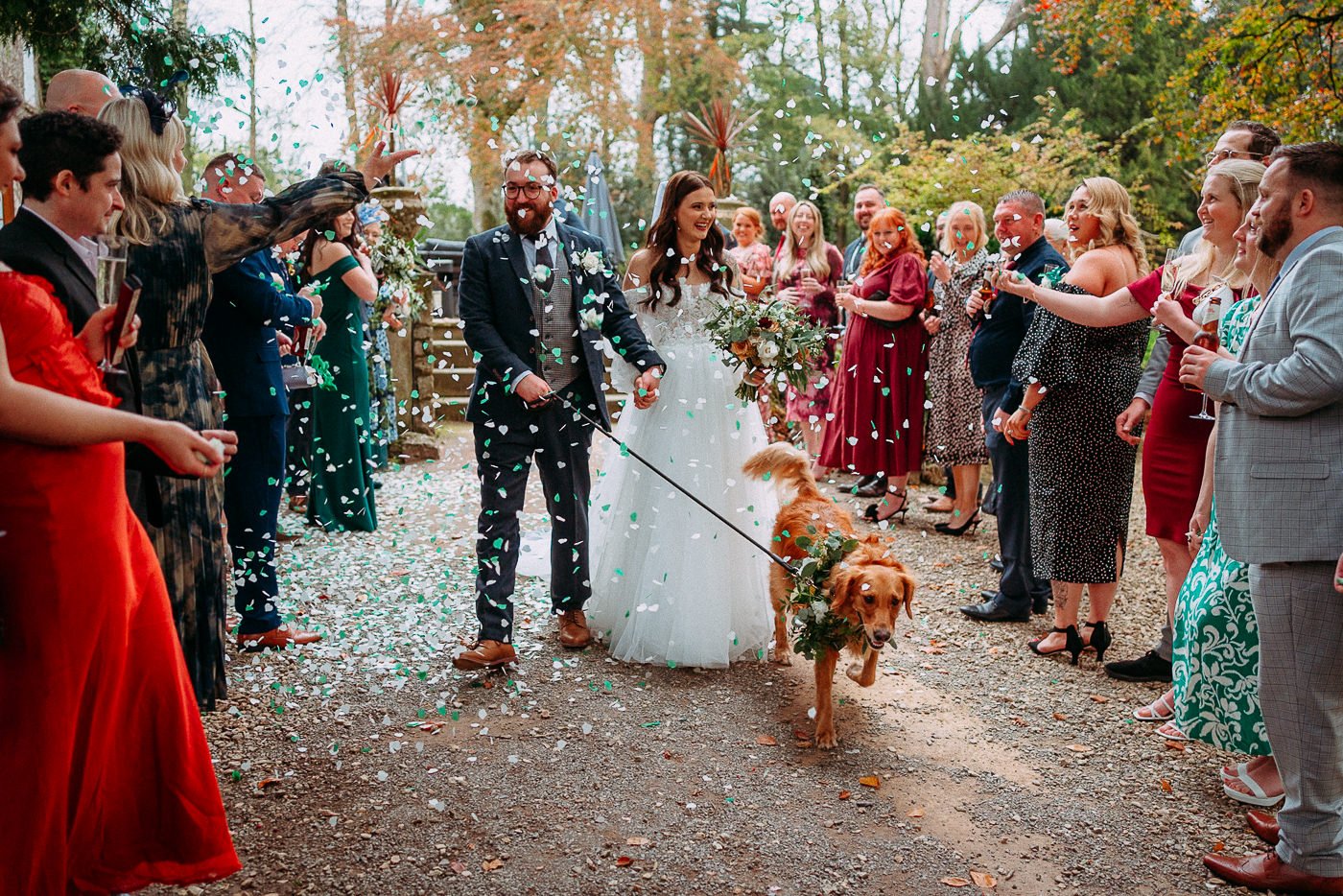 A wedding celebration outdoors with a bride and groom walking through a confetti shower, surrounded by guests on either side, in an autumn setting.
