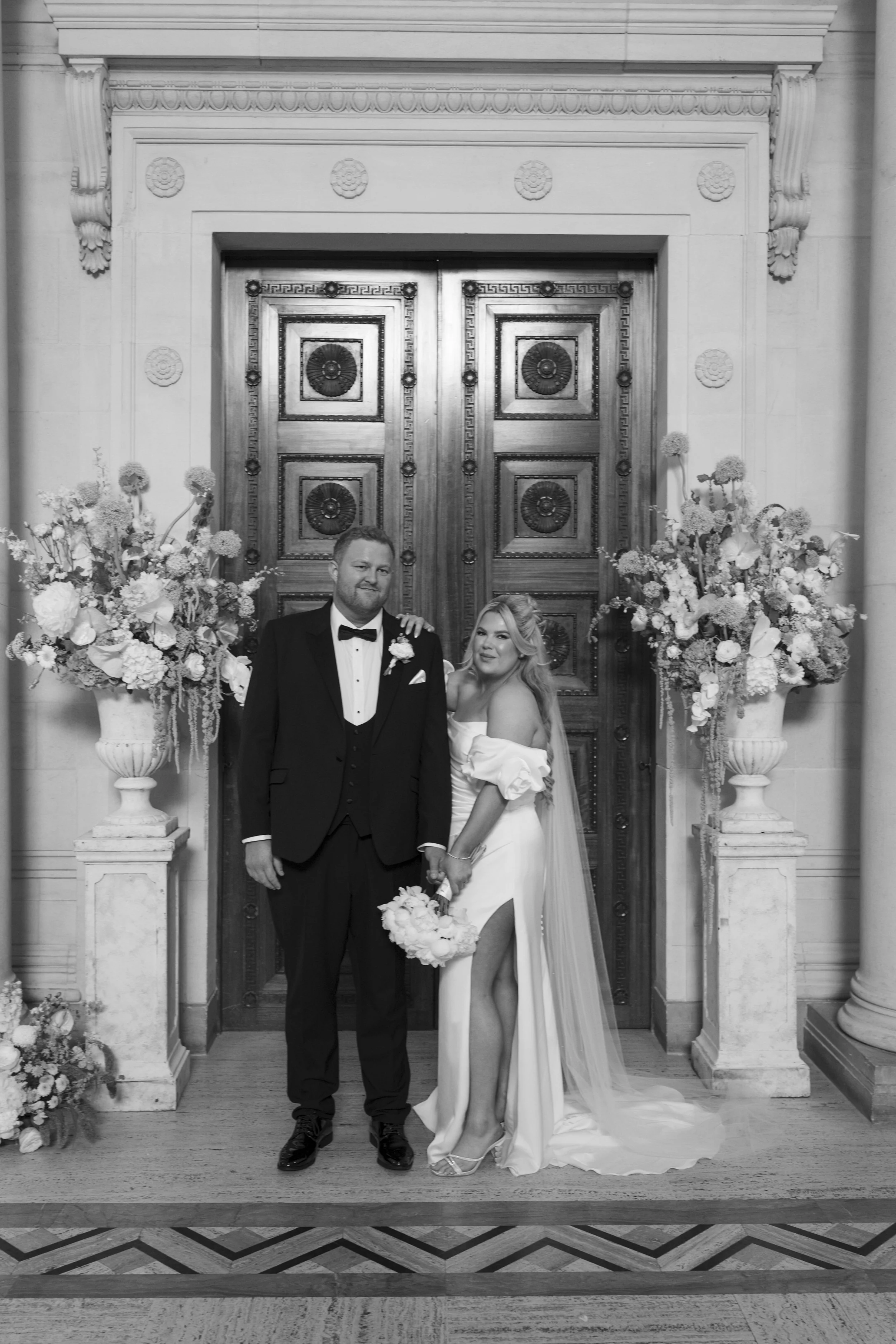 Black and white photo of a bride and groom standing in front of a large wooden door decorated for a wedding, surrounded by flower arrangements.