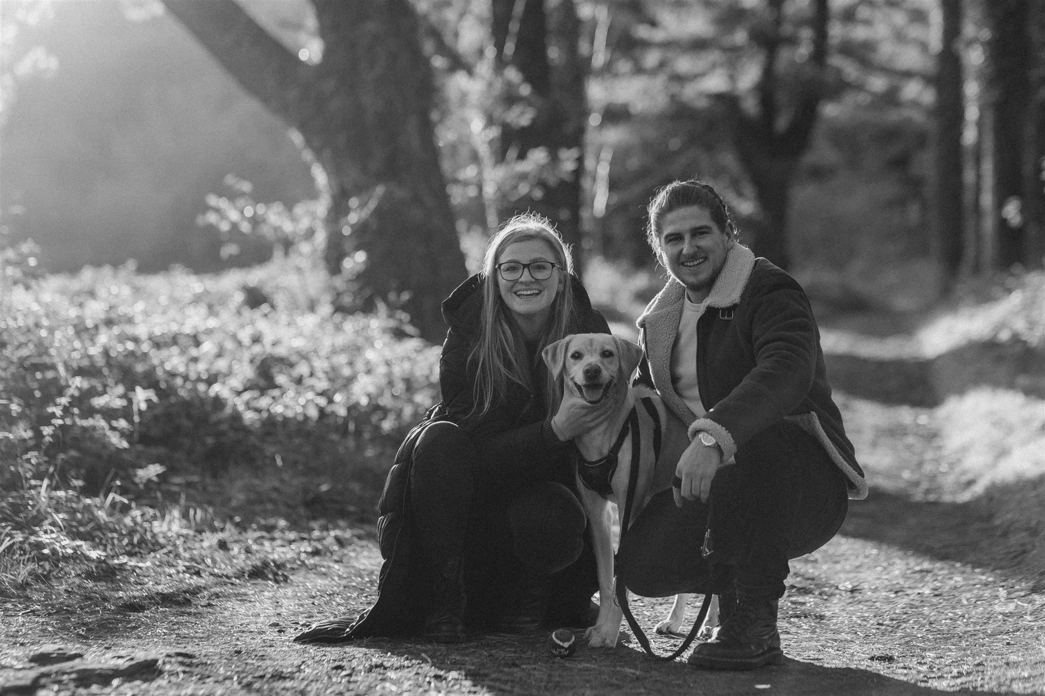 A black and white photo of a smiling couple with a happy dog on a leash, in a forest with sunlight filtering through trees.