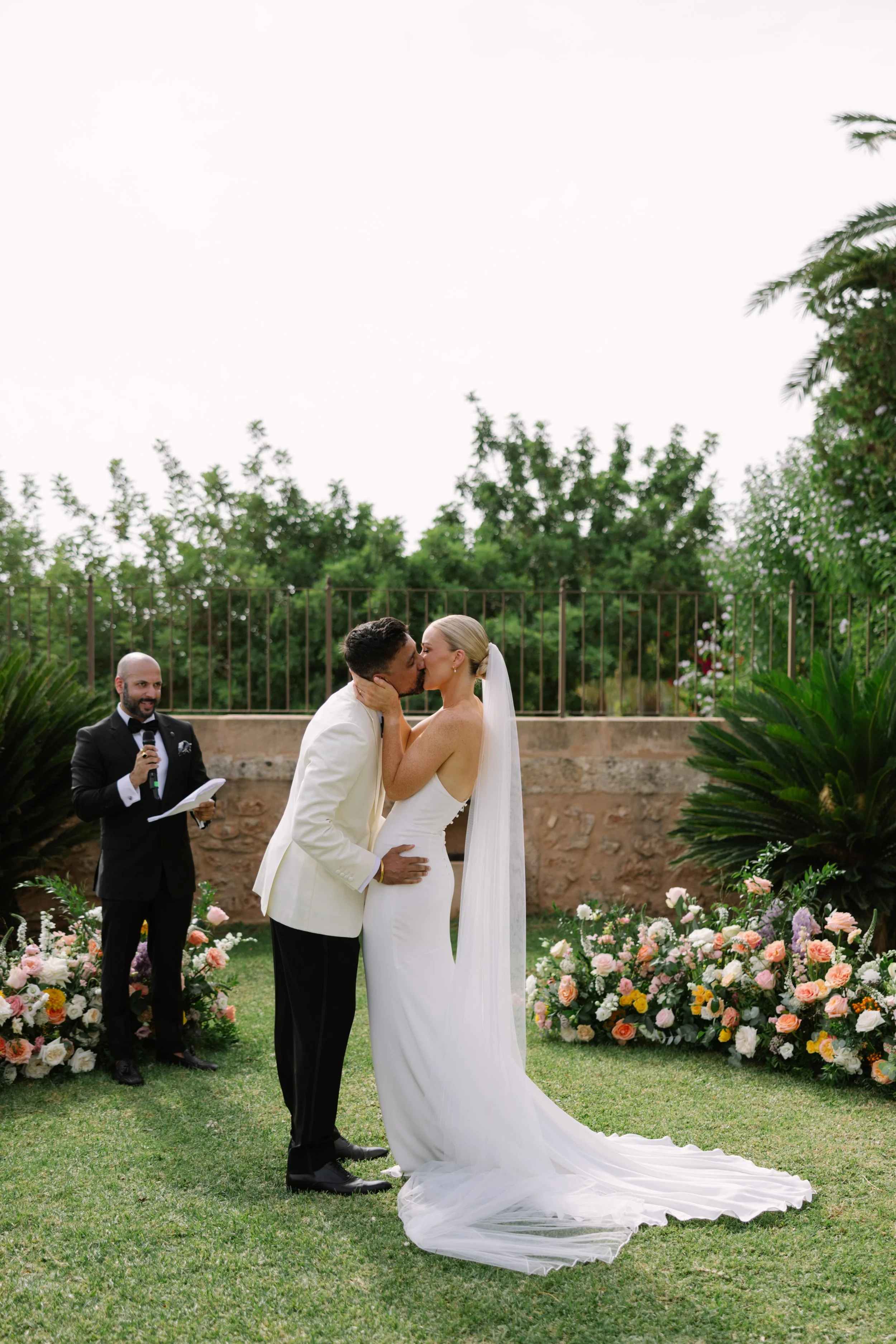 A bride and groom kiss during their outdoor wedding ceremony, with an officiant holding a microphone and papers watching in the background, surrounded by floral arrangements.