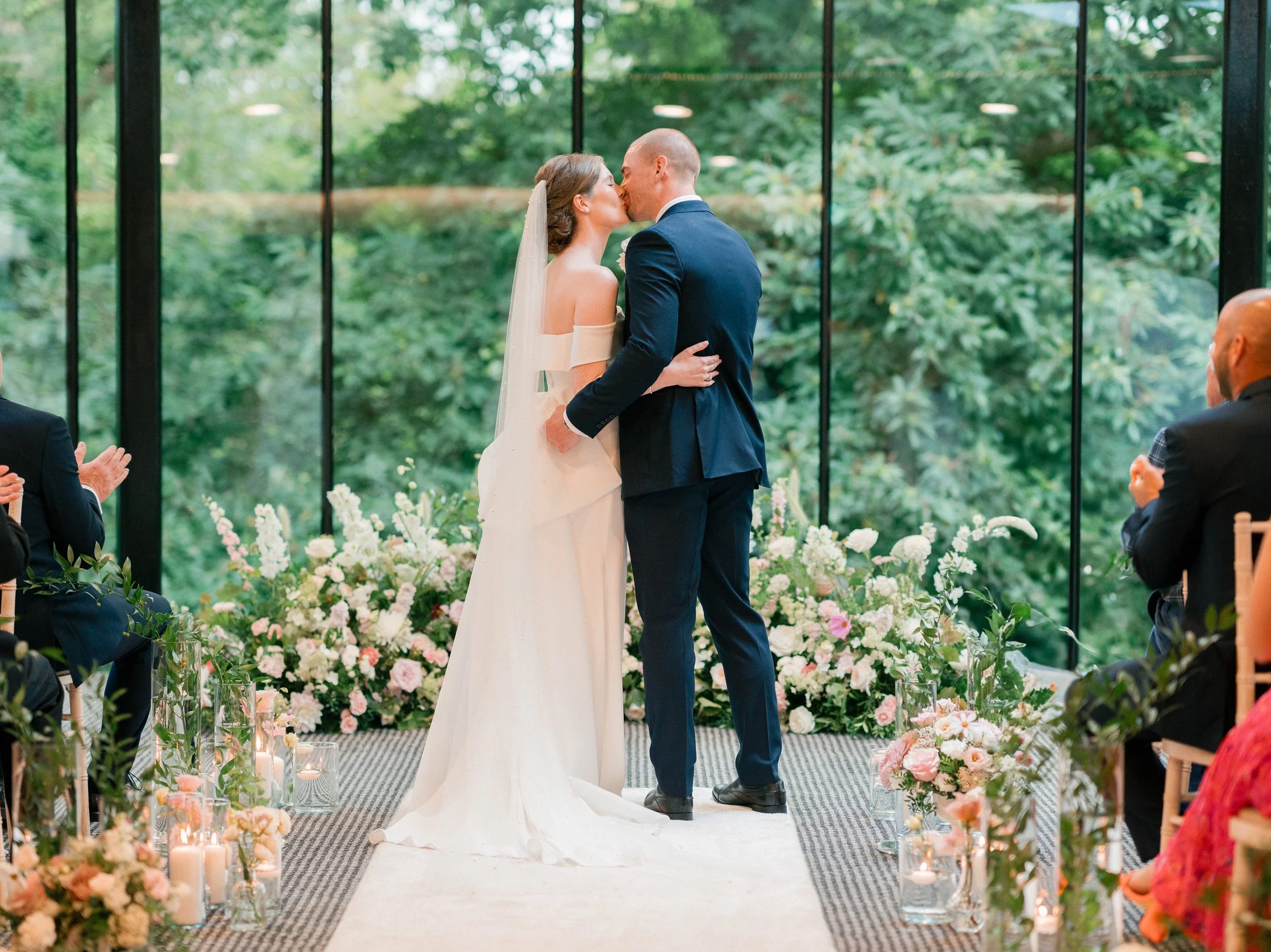 A bride and groom kiss during their wedding ceremony in front of large windows with green trees outside. The scene is decorated with pink and white flowers and candles, and guests are clapping.