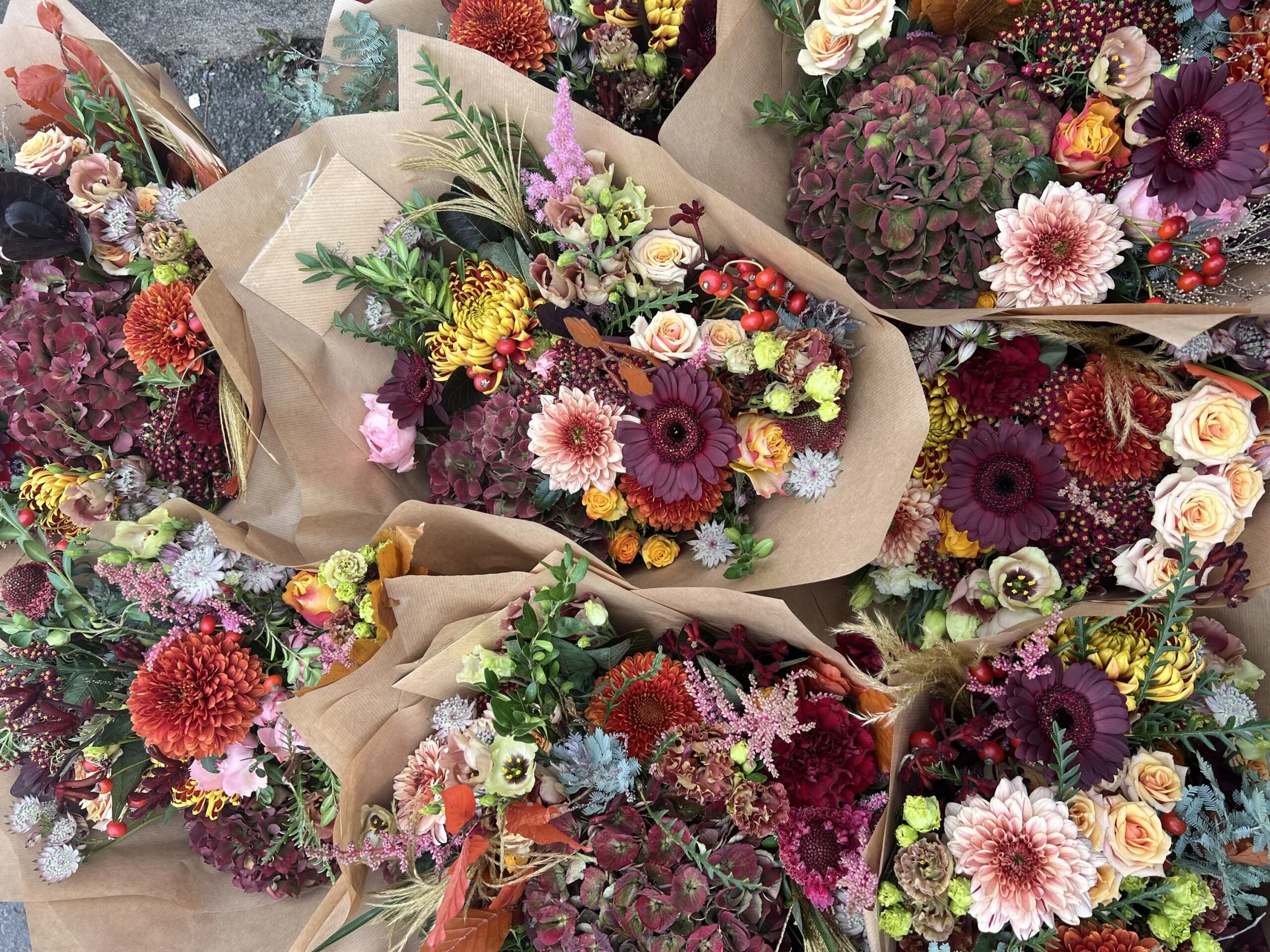 multiple gift bouquets in autumnal tones, pinks, oranges and reds all wrapped in brown kraft paper. flowers include hydrangea, gerberas and roses