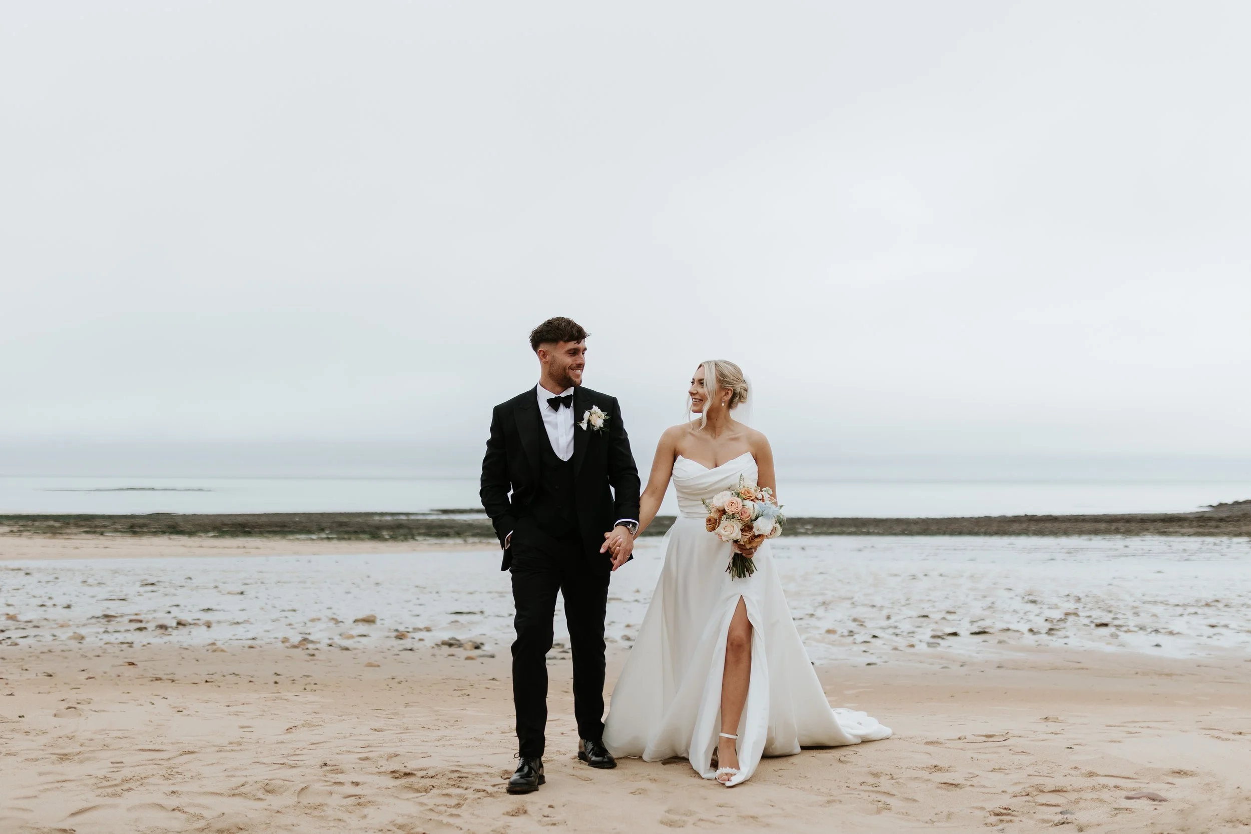 A bride and groom walking hand in hand on a sandy beach, both smiling, with an overcast sky in the background.