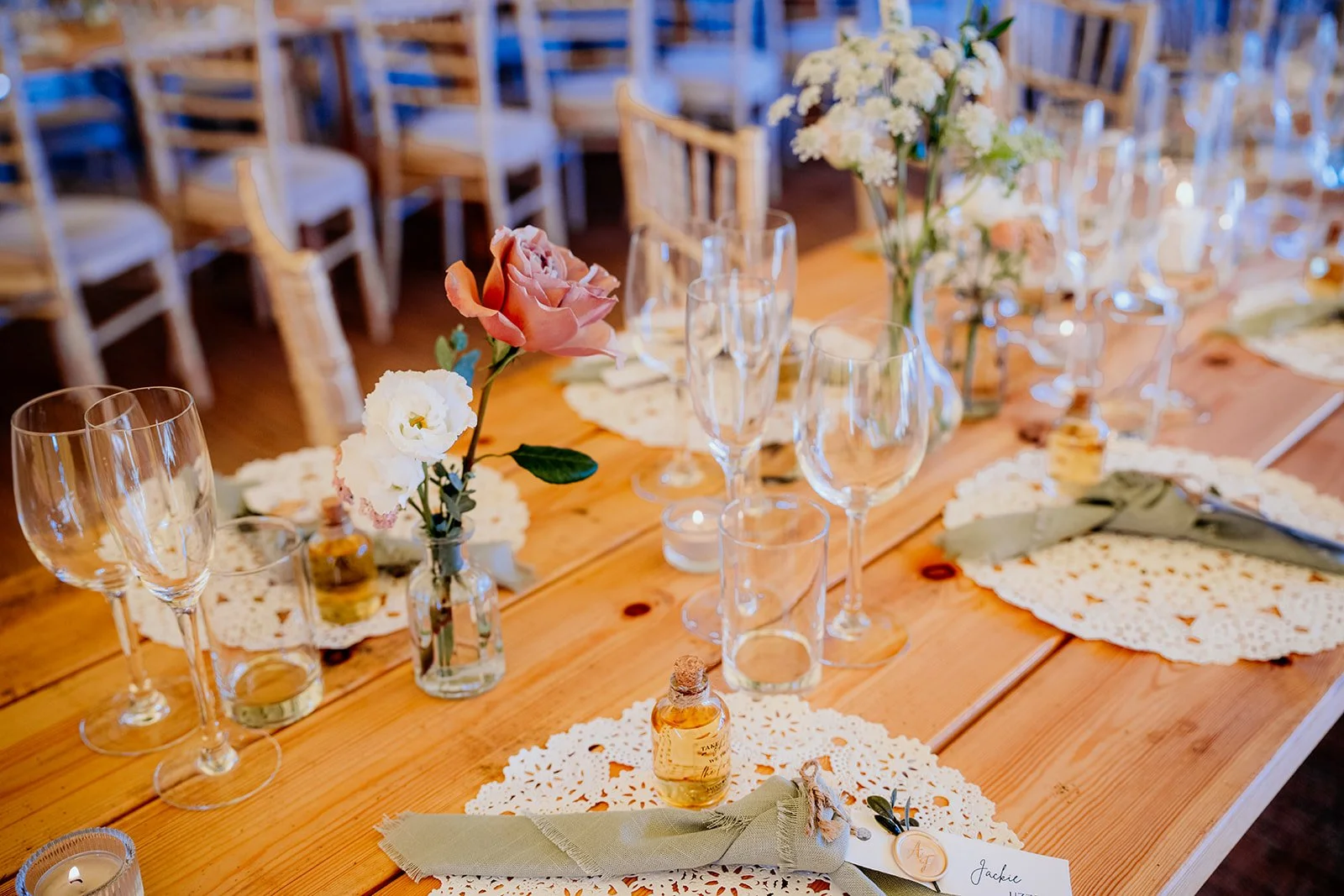 wooden tables with pink rose in a small vase with other white flowers. napkins with place names on, candles and glassware lining the table. 