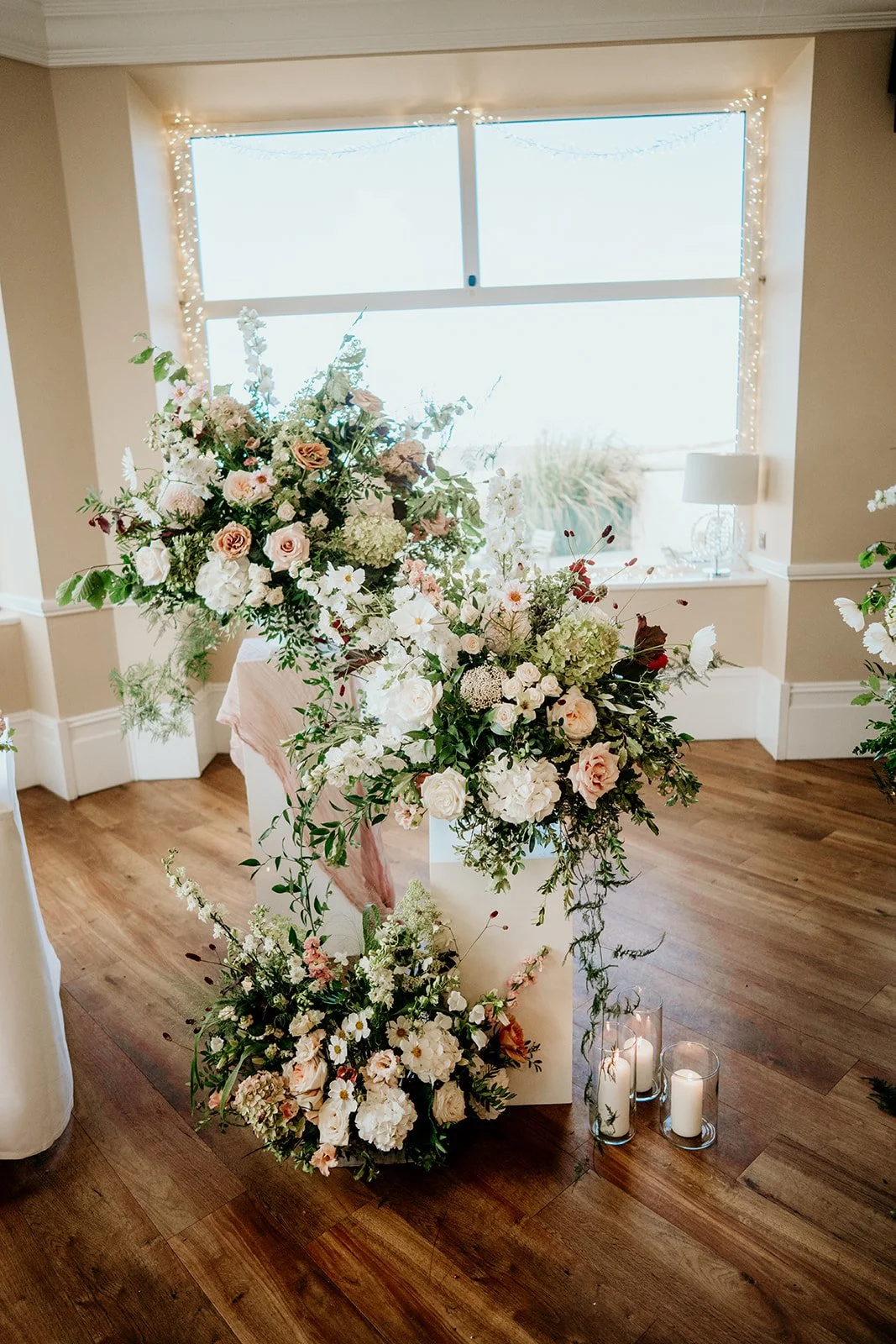 Large display of flowers at an indoor wedding ceremony. Fabric draped over white wooden plinths. Three large flower arrangements with flowers including hydrangeas, roses and lots of greenery and white and nude coloured flowers . all arrangements plac