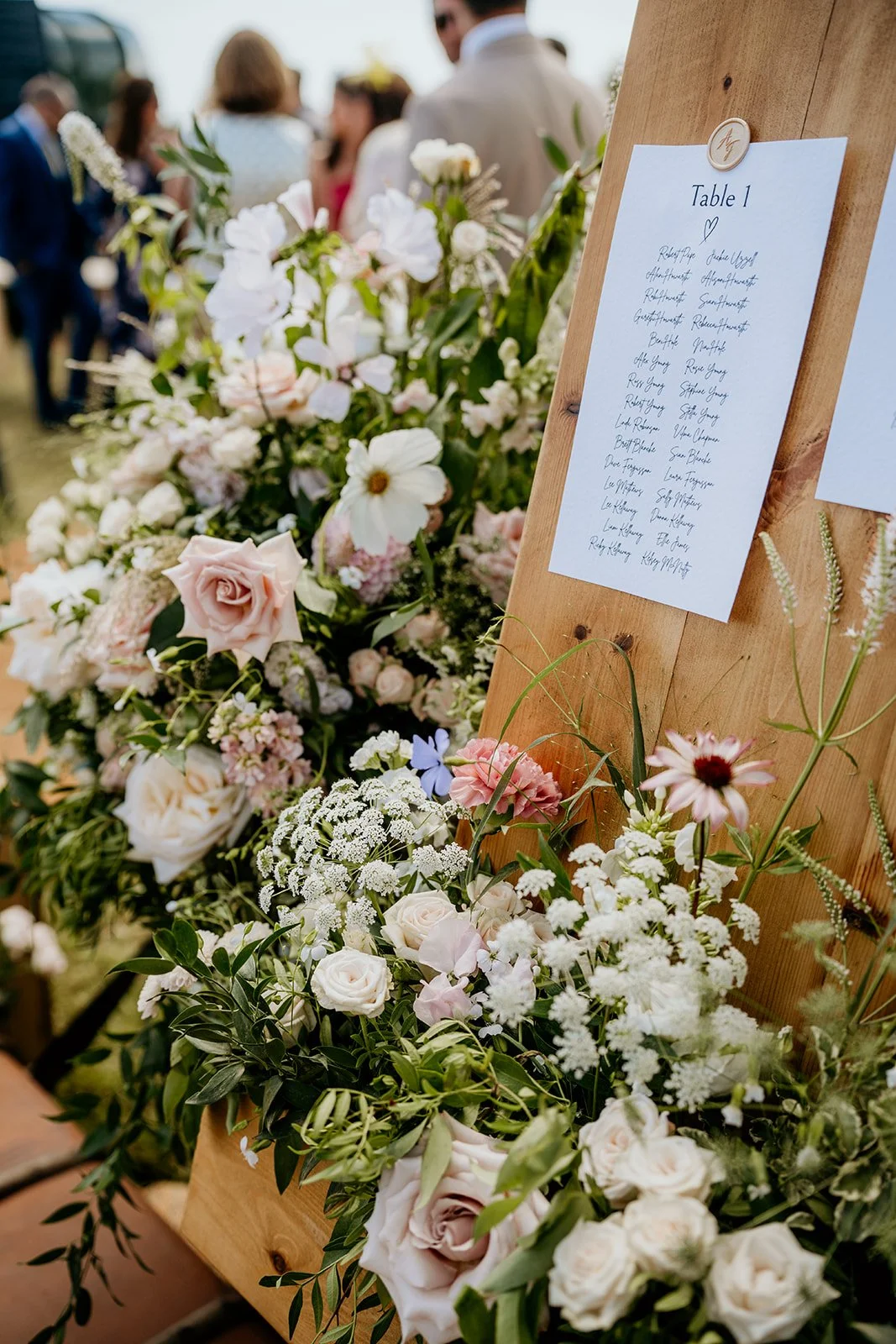 A wedding seating chart on a wooden stand, surrounded by a large floral arrangement with white, pink, and purple flowers, at an outdoor wedding reception with guests in the background.