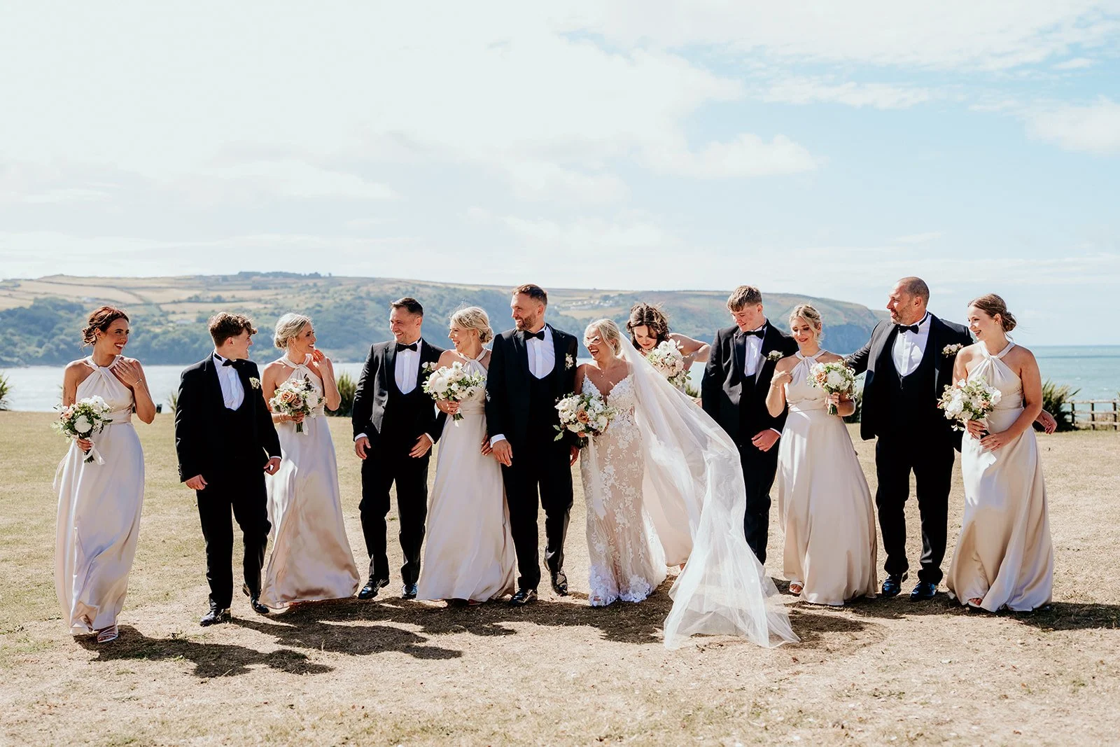 A wedding party outdoors on a grassy field with a ocean view in the background, including a bride, groom, and several bridesmaids and groomsmen, all smiling and holding bouquets.