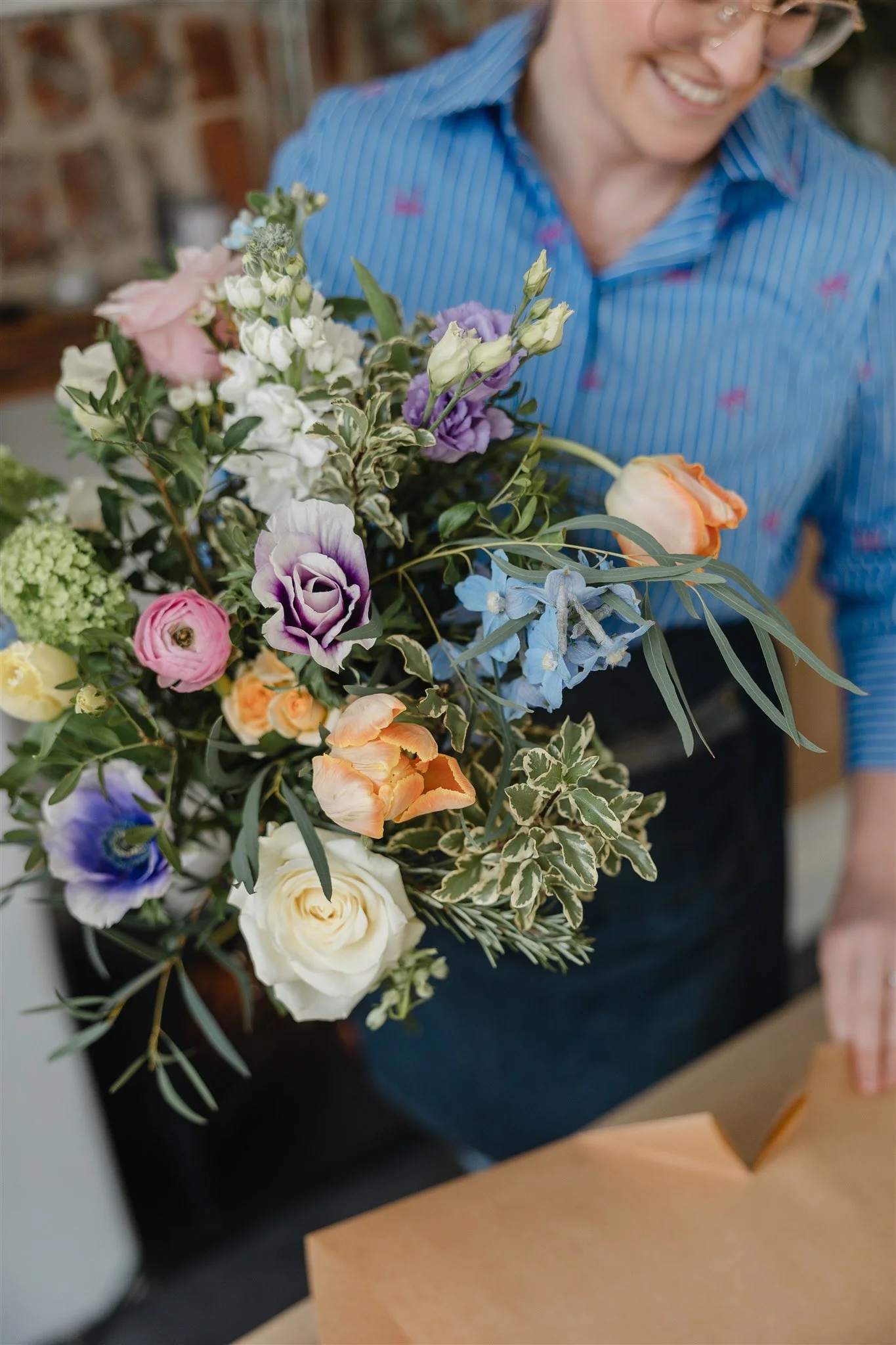 a woman in glasses smiling, in a blue patterned shirt and denim apron holding a very colourful flower bouquet, with cut kraft paper ready to gift wrap the bouquet an exposed brick wall in the background.