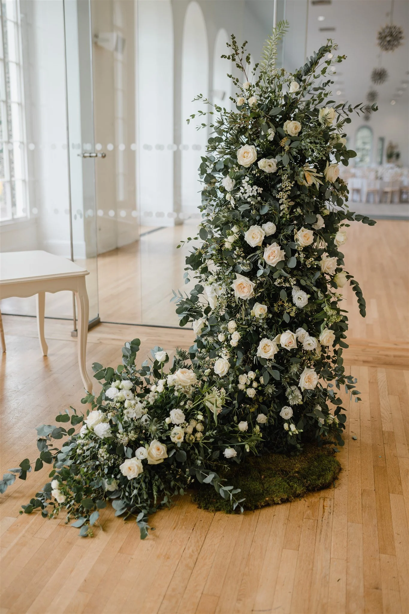 A tall floral arrangement with white roses and greenery, arranged in a cascading style, placed indoors on a wooden floor.