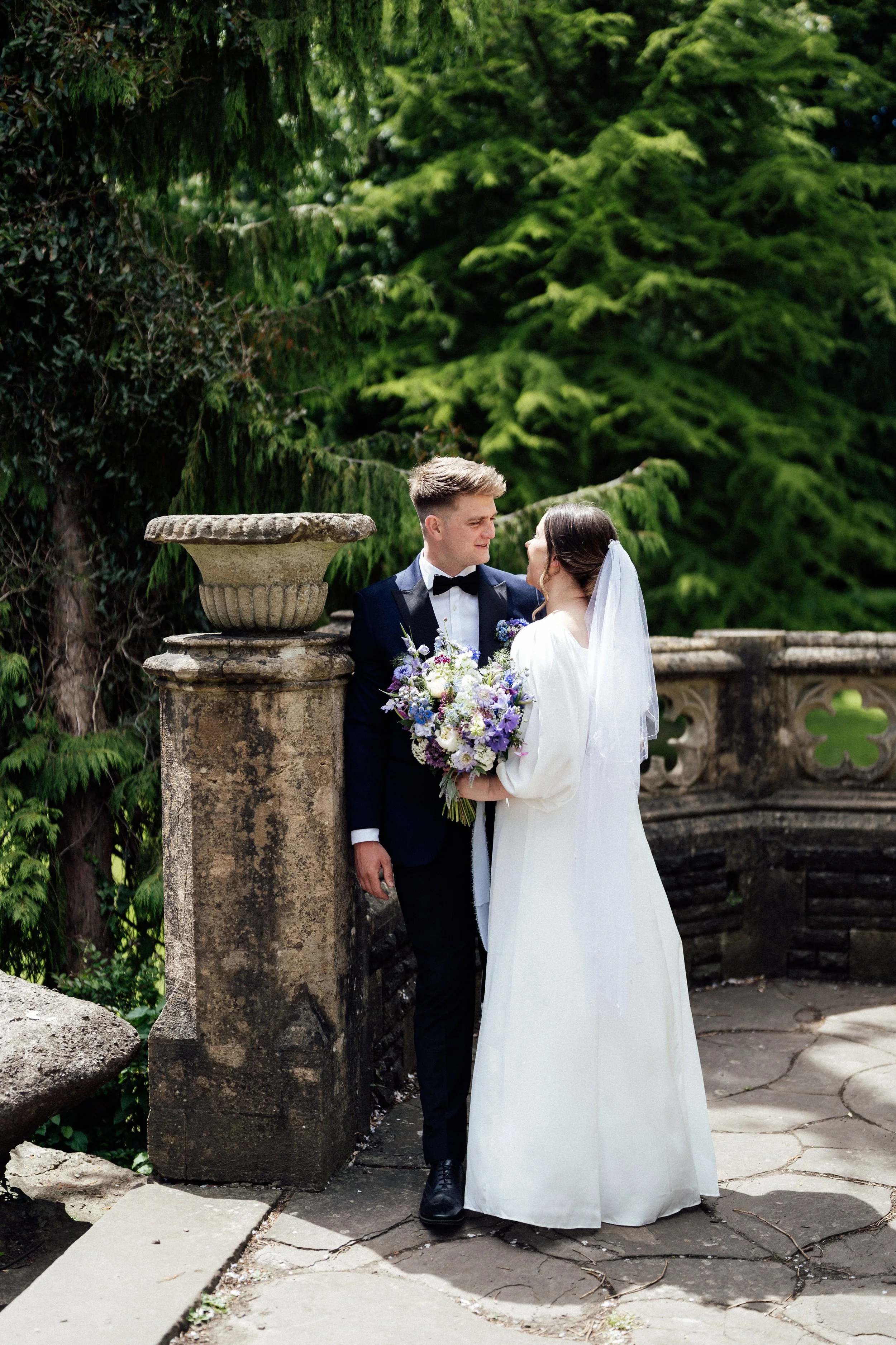 Wedding couple outdoors, bride holding a bouquet, bride in white dress with veil, groom in black tuxedo, lush green trees background, stone balustrade.