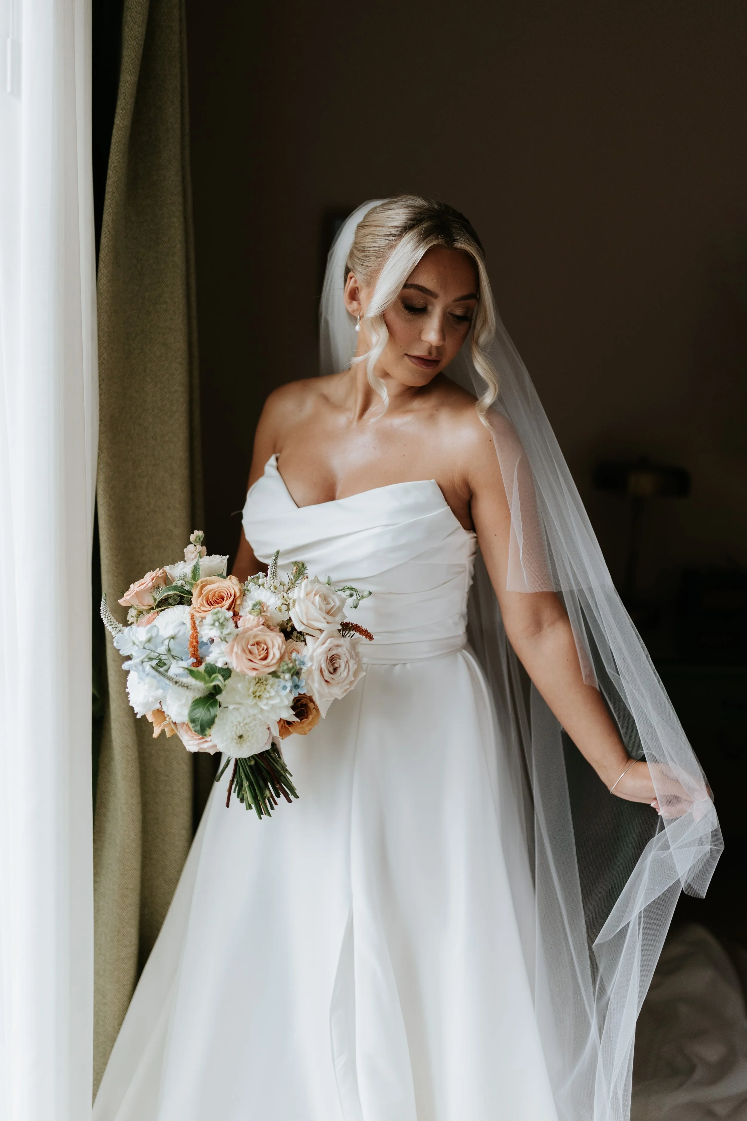 A bride in a white wedding dress holding a bouquet of pink and white flowers, standing near a window with curtains, looking down.