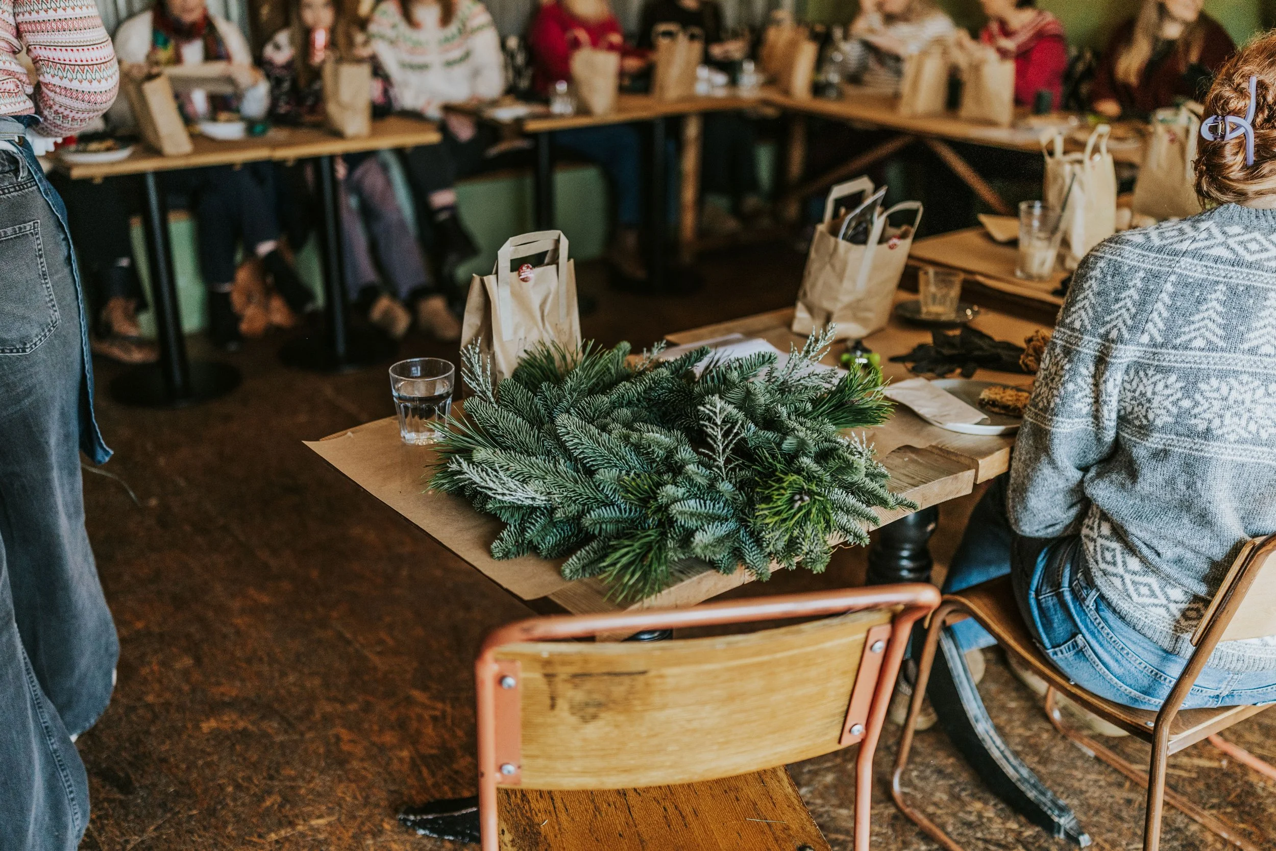 Indoor gathering with people sitting around tables decorated with a Christmas greenery centerpiece, possibly for a holiday event.