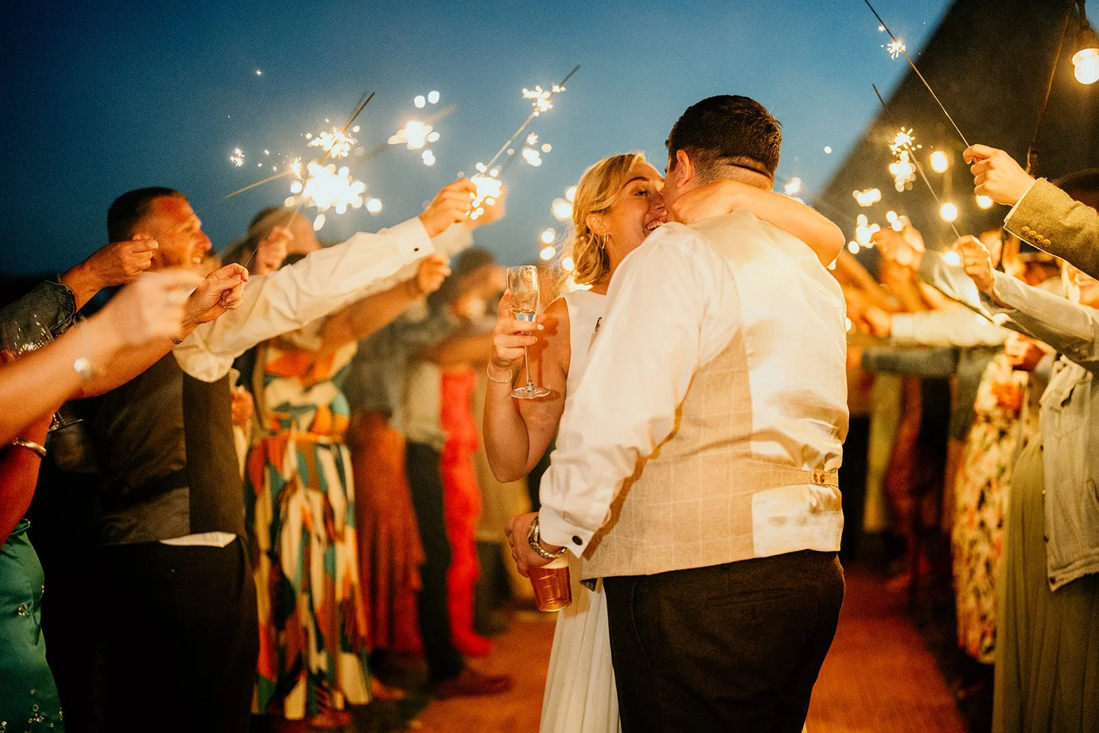 a bride and groom kissing in a line of their guests holding sparklers above their heads. a tipi in the background, the night sky is dark blue but softly lit by the sparklers.