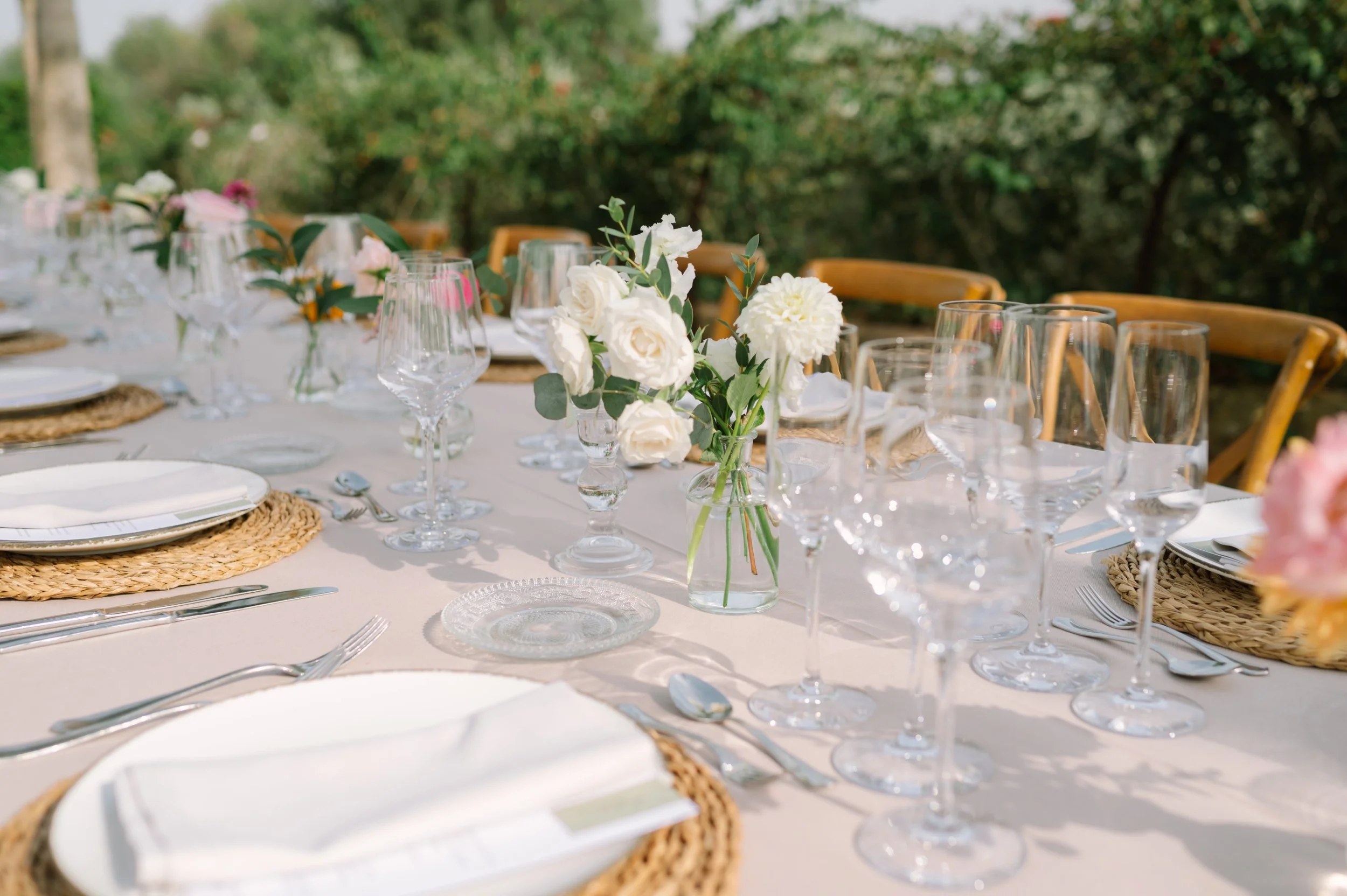 A beautifully set outdoor dining table with glassware, white plates, and floral centerpieces, surrounded by wooden chairs in a garden setting.