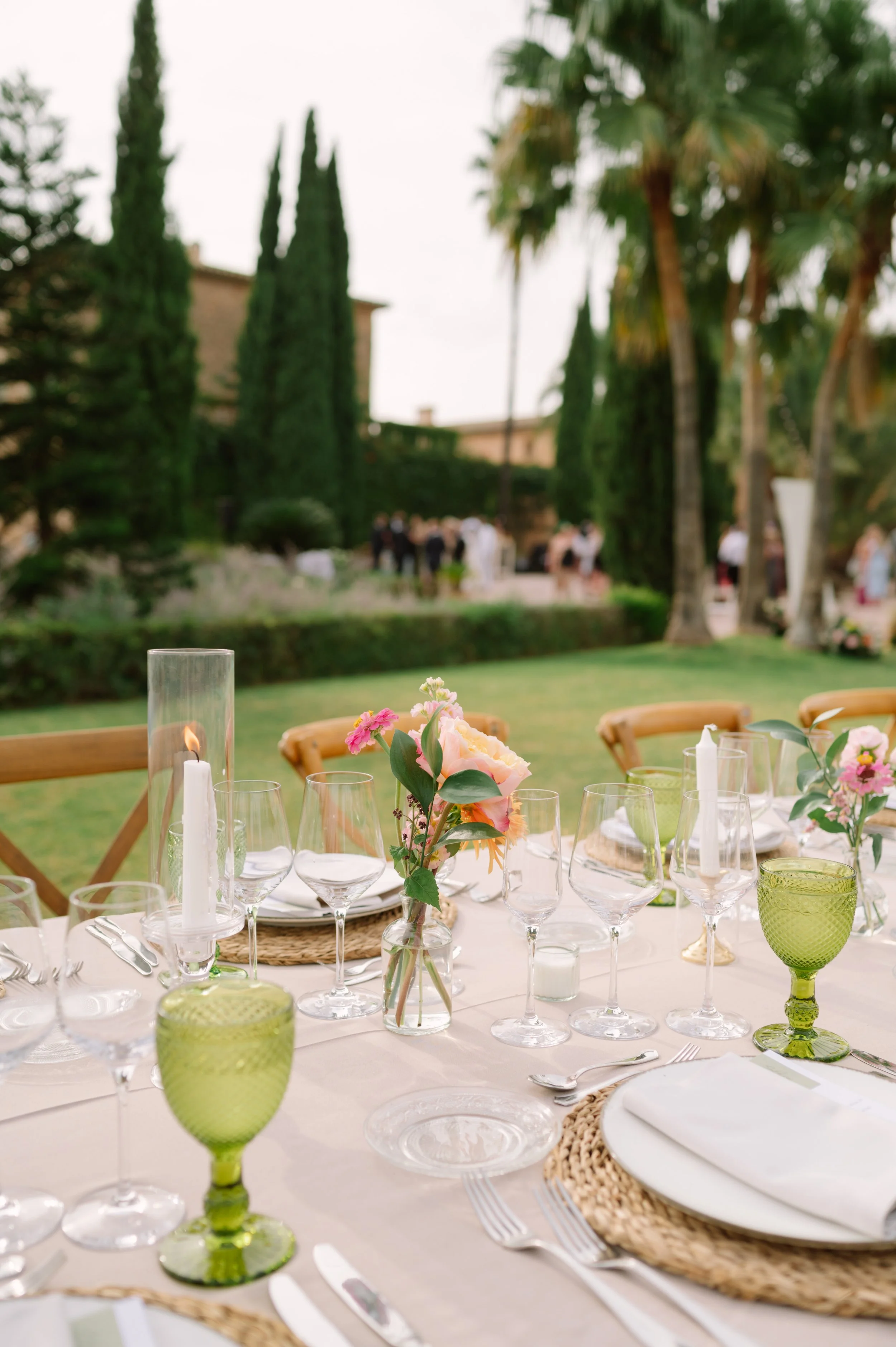Elegant outdoor table setting with glassware, flowers, and candles, surrounded by tall trees and greenery.