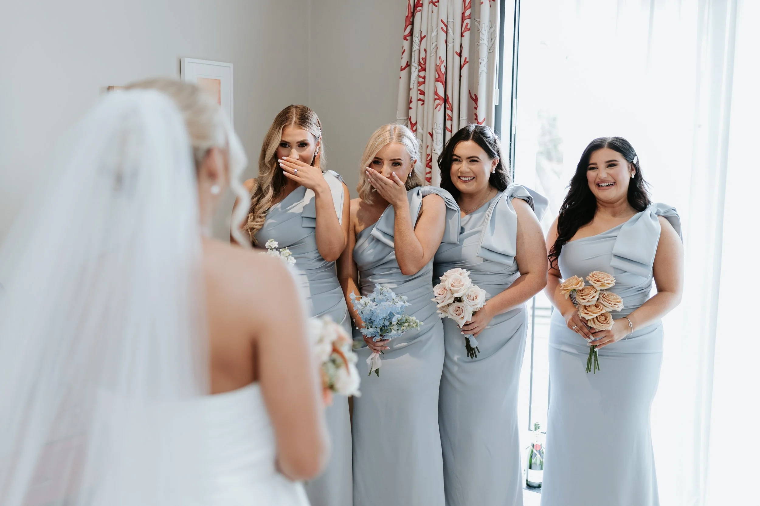 Bridesmaids in matching light blue dresses reacting with smiles and surprise as the bride, out of focus, stands in front of them holding a bouquet.