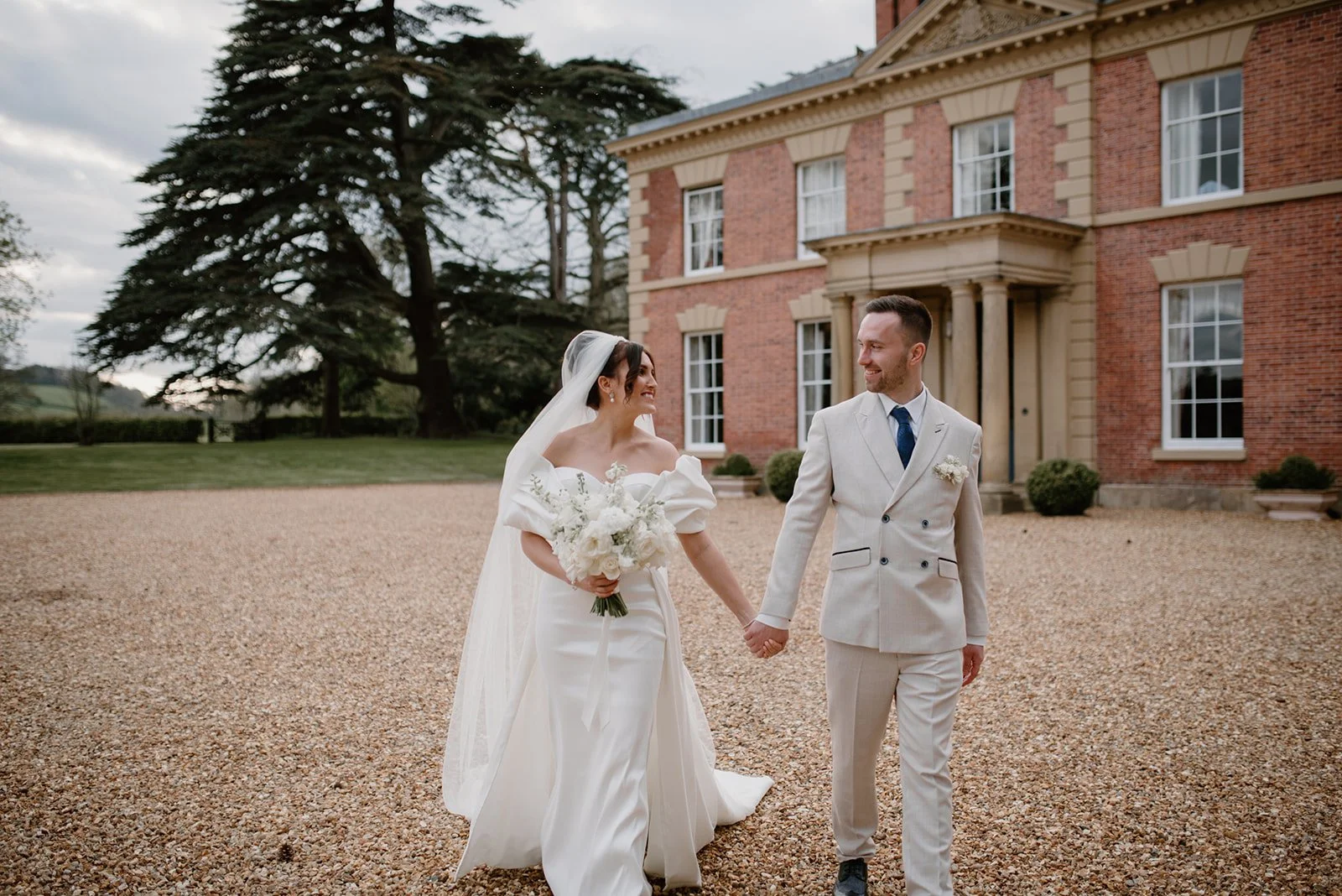 A bride and groom walking hand in hand outside a large brick house with greenery in the background.