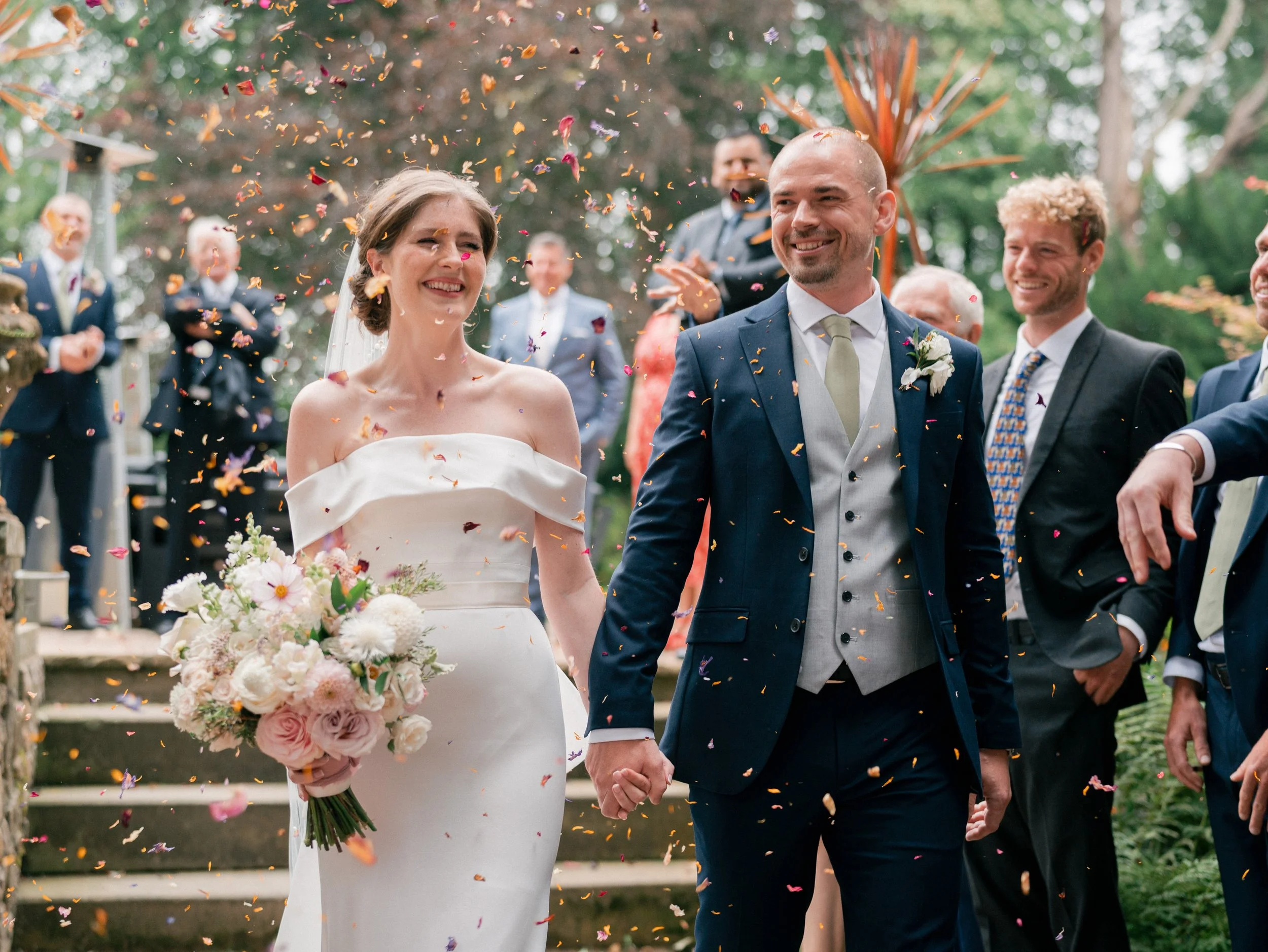 A bride and groom holding hands and smiling, surrounded by guests, with confetti falling during their outdoor wedding celebration.