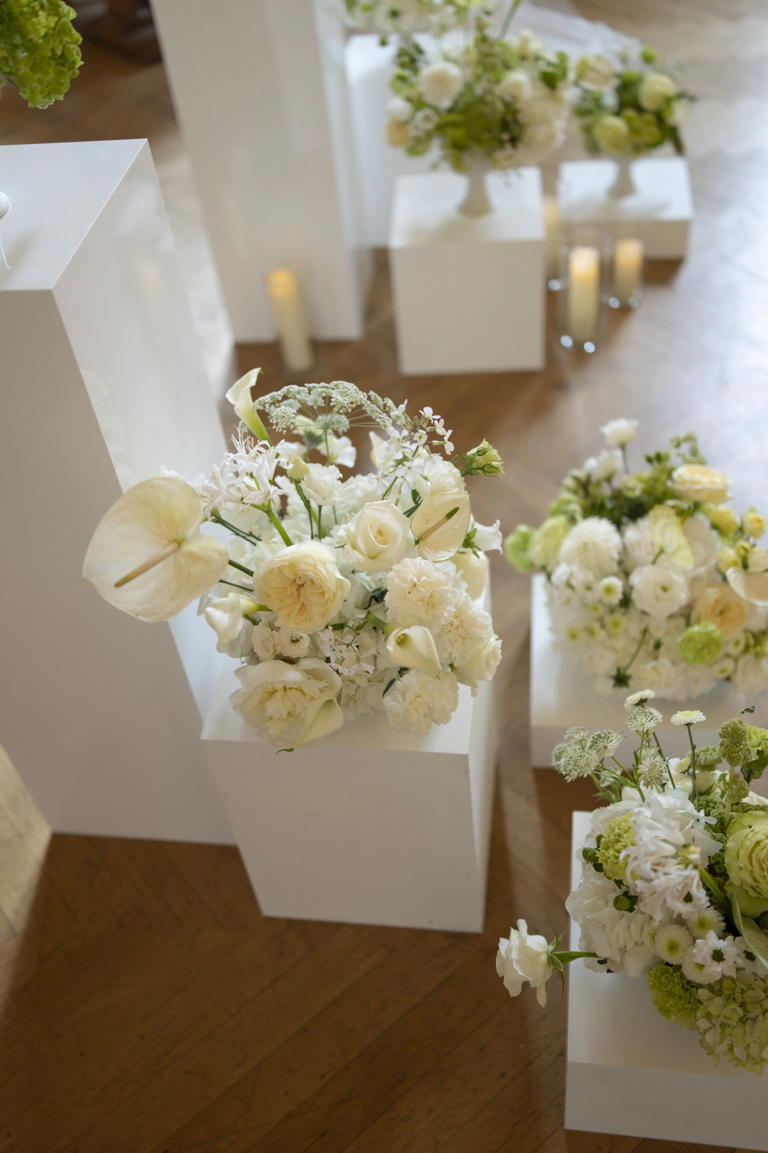 White and green flower arrangements placed on white wooden plinths all displayed at different heights, candles in the background with focus on the flowers in the foreground against dark wooden floor. 