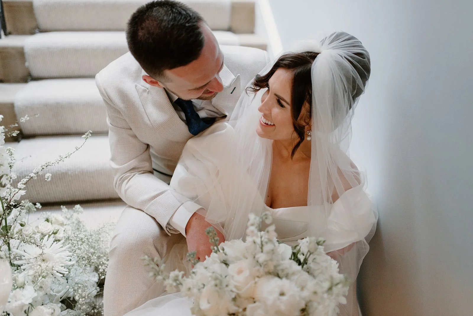 A bride and groom smiling and looking at each other, with the bride sitting on stairs holding a bouquet of white flowers, and the groom leaning over her, in a wedding setting.