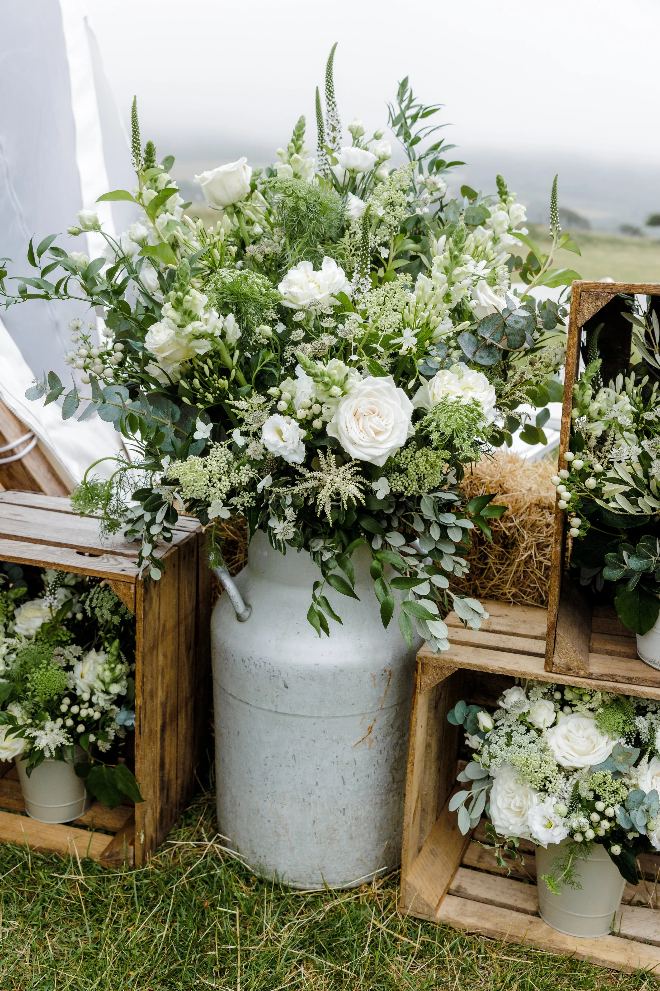 White flower arrangement in a white pitcher, surrounded by wooden crates with additional small floral arrangements, set outdoors on grass.