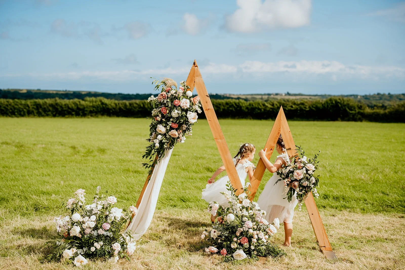 an outdoor ceremony set up with triangle shapes covered in pink and white flowers and some draped cream fabric in a field, 2 little girls in white dresses are playing in the background amongst the flowers in the field.