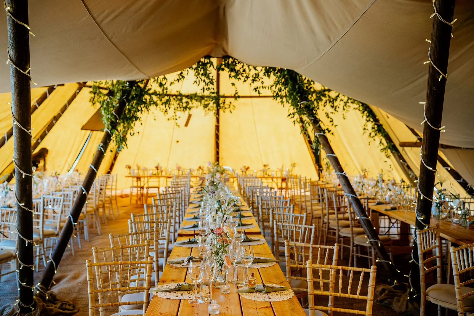 inside of a tipi set up for a wedding reception. long wooden tables with chairs either side, decorated with glasses, bud vases and candles. Green vines and foliage in the roof of the tipi shown in the background. fairy lights wrapped around the tipi 