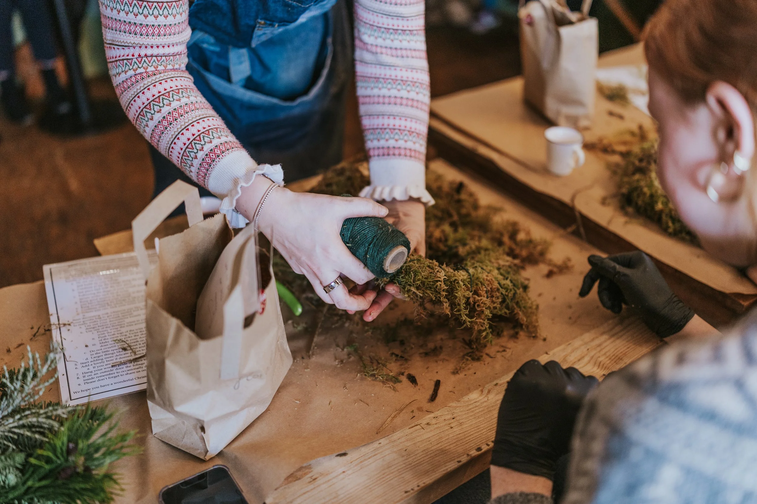 Two women working on creating a botanical arrangement with moss and foliage at a wooden table, with craft supplies and paper bags nearby.