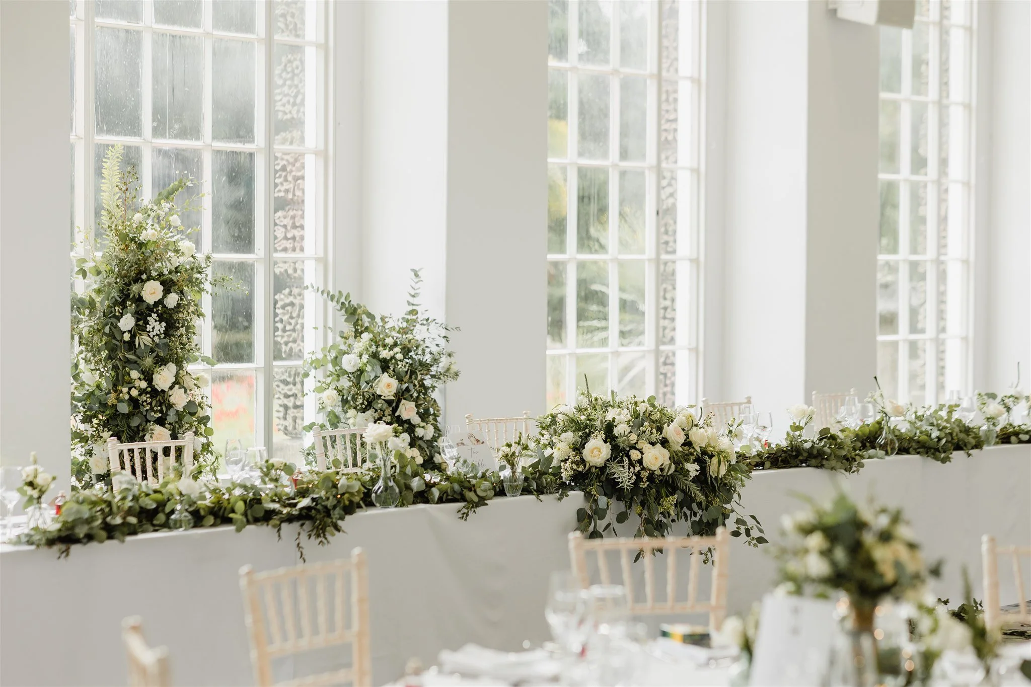 a grand room with floor to ceiling windows, decorated with white flowers and an abundance of greenery. 