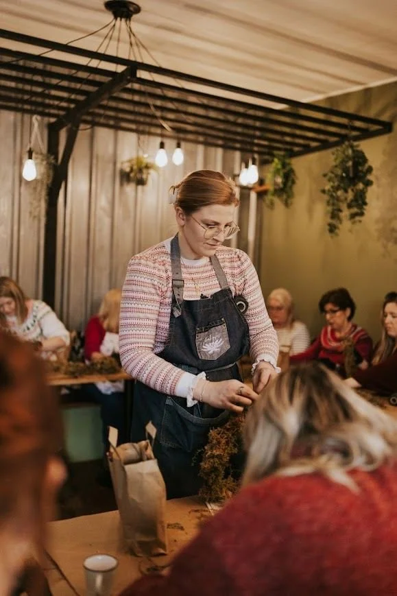 A women in a stripey red Christmas jumper with a denim aproteaching a christmas wreath making workshop in a cozy coffee shop space. the florist is tying a knot in the moss wreath ring base so the participant can start adding foliage to their wreath.
