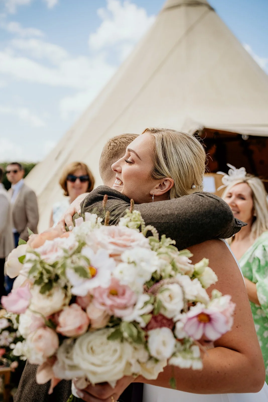 A bride hugging a guest at an outdoor wedding, holding a bouquet of pink and white flowers, with a beige tent and other guests in the background.